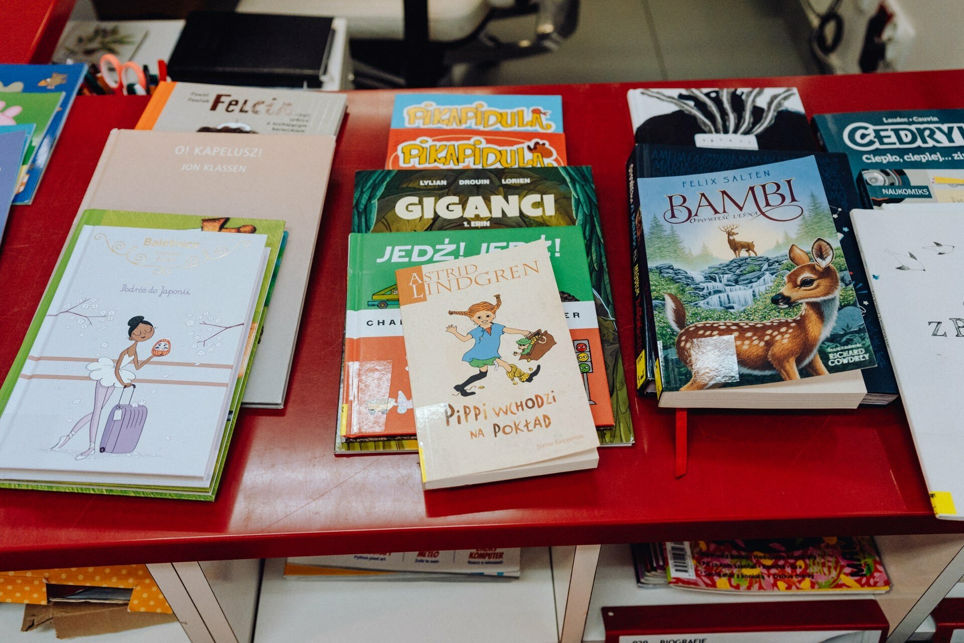 Exhibition of children's books on the red table. Titles include "Pippi Comes on Board," "Bambi" and "Giants." The colorful covers feature fun illustrations and characters, such as a girl with a suitcase and a deer. Captured by a photographer from Warsaw, some of the books are stacked.   