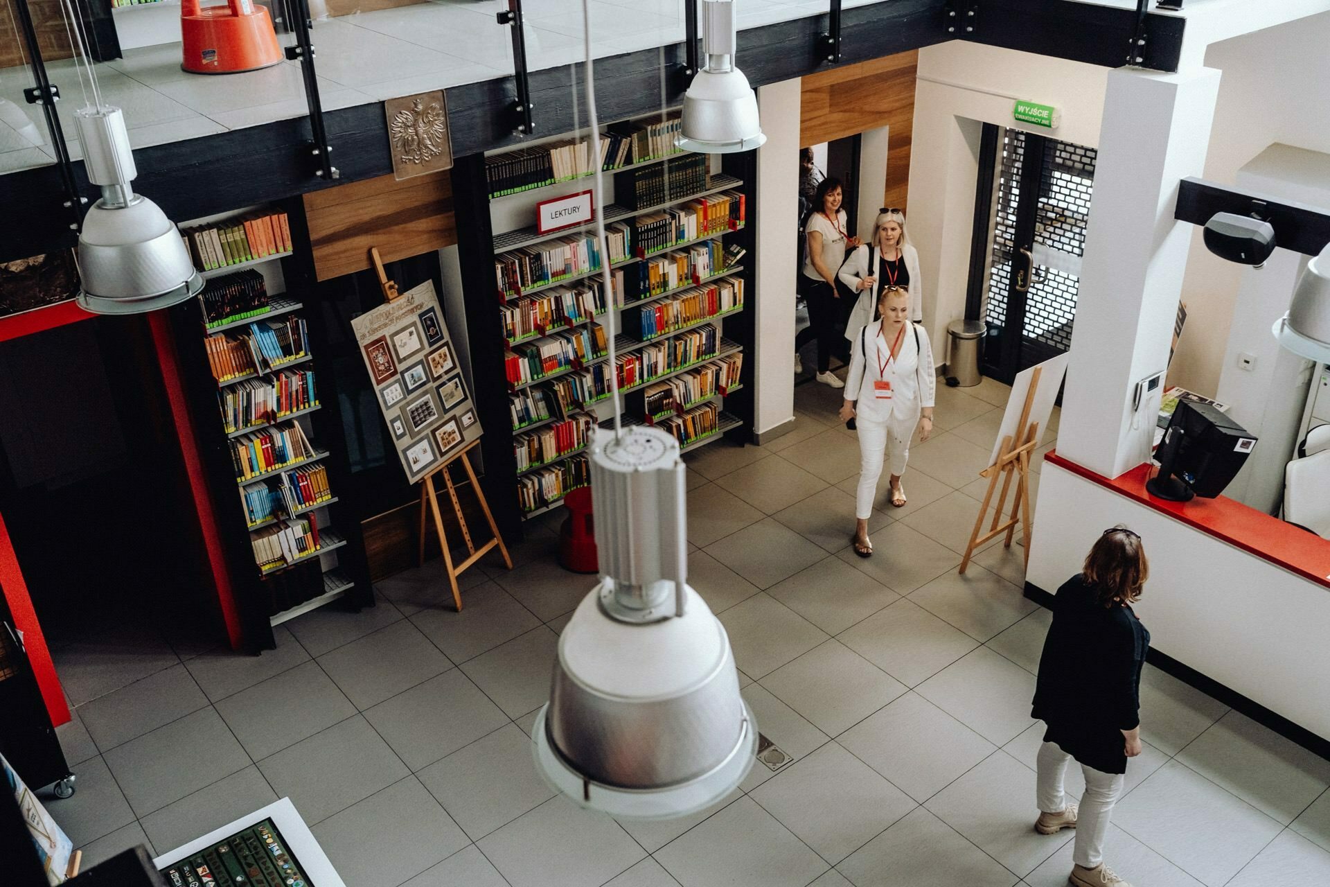 A brightly lit library with shelves filled with colorful books. Several people walk or stand, immersed in conversation. The space has high ceilings with modern pendant lights, and several easels hold photos or artwork by the latest event photographer Warsaw.  