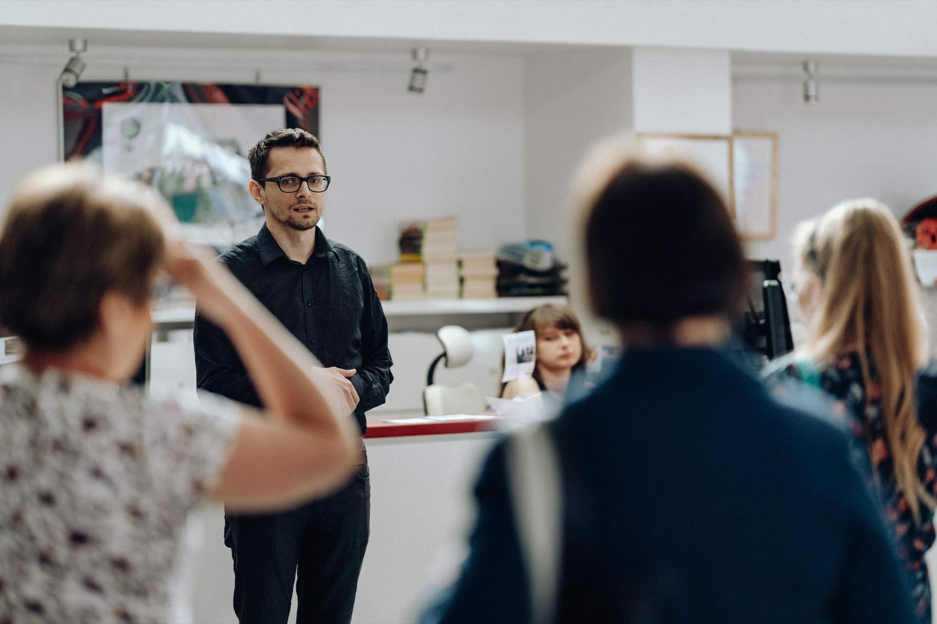 A man in a black shirt is standing in front of a group of people who are listening intently. He appears to be in a public space, such as an office or community center. In the background are books on shelves and a woman sitting at a desk, brilliantly captured by event photographer Warsaw.  