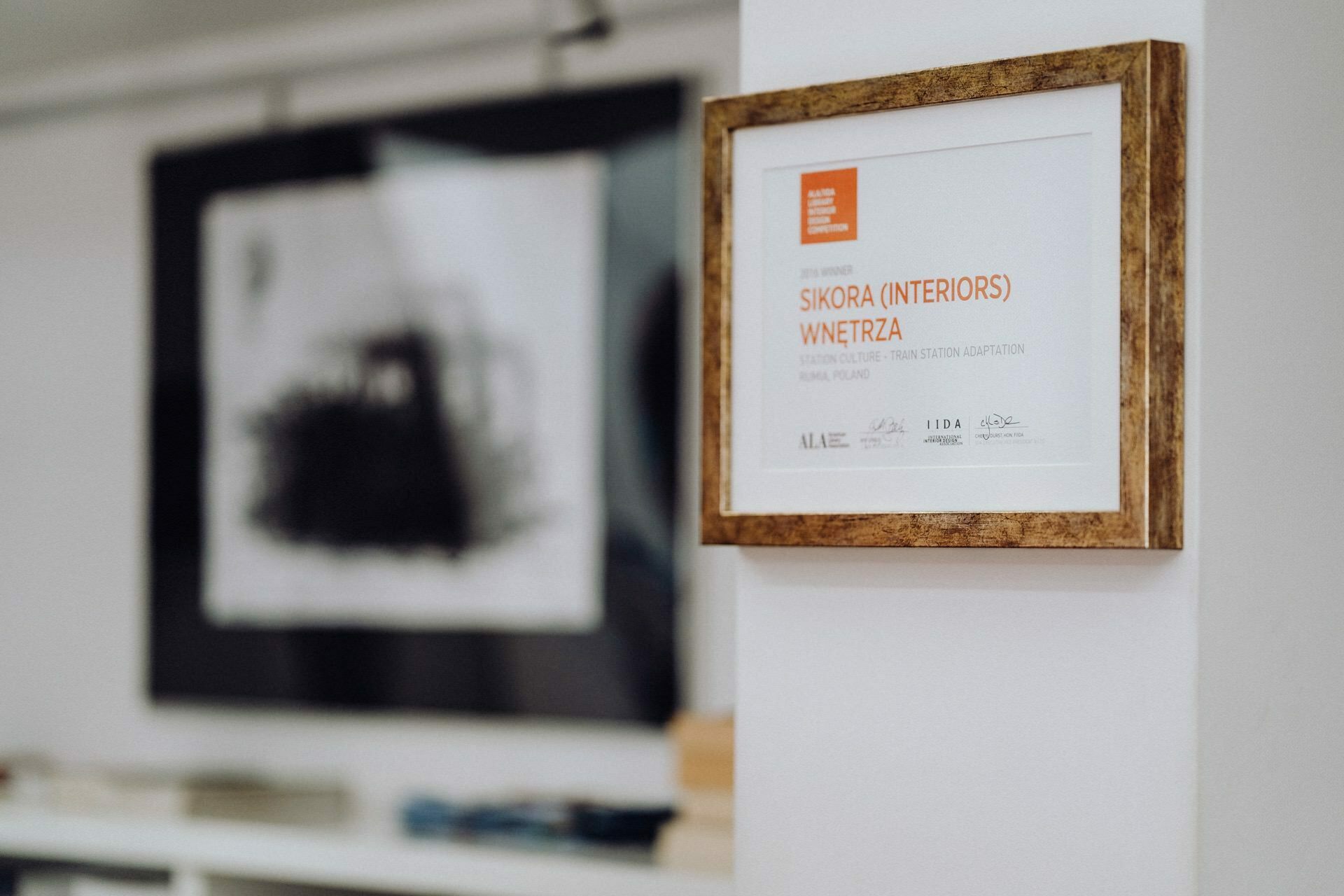 On the white wall hangs a certificate in a wooden frame, awarded to "Sikora (Interiors) Werza" for cultural and translation adaptation in Krakow. The black and white graphics and white shelves of objects were captured by a photographer from Warsaw on a blurry background. 