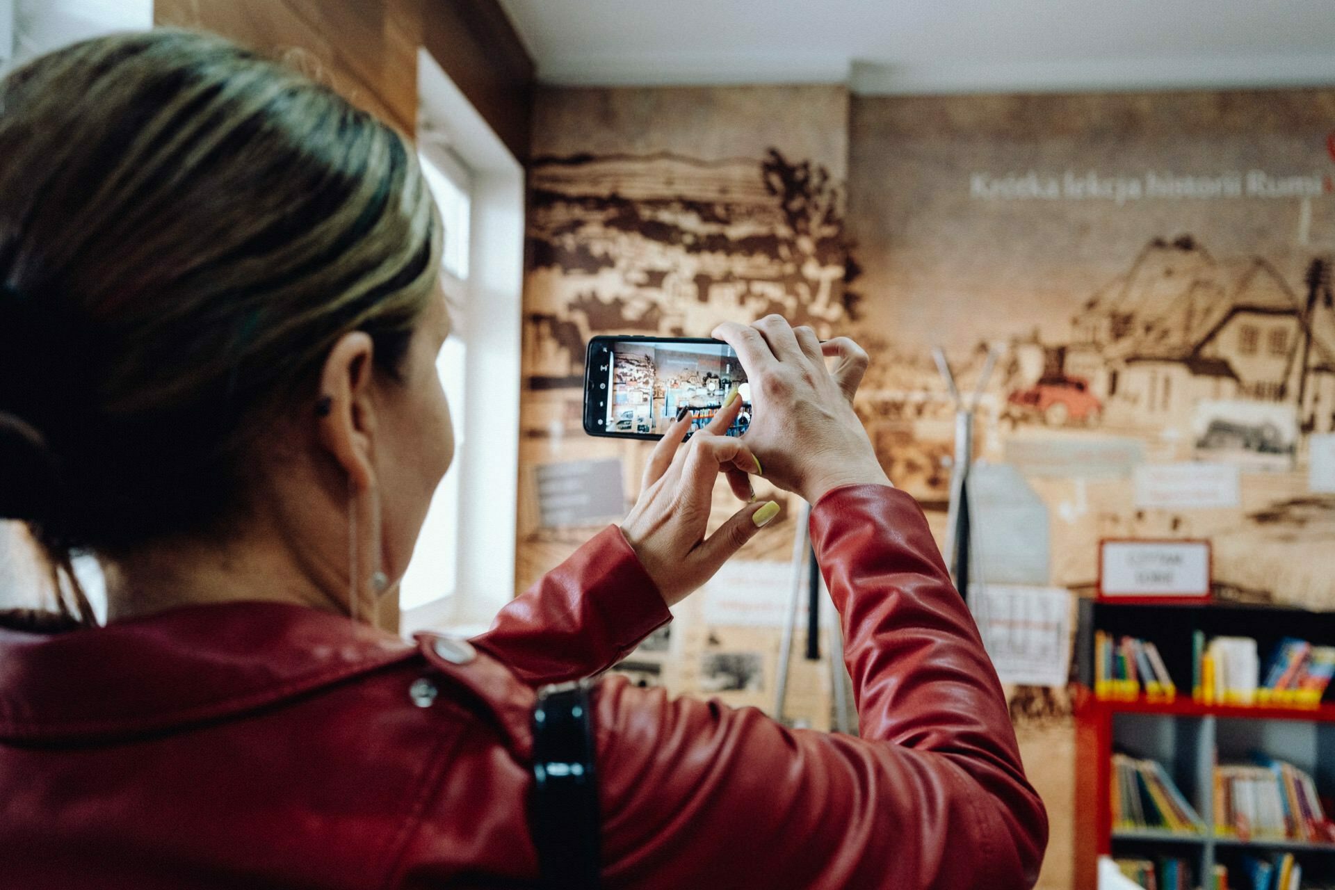 A woman dressed in a red jacket takes a photo of a museum exhibit with her smartphone. The exhibit features historical images and texts on the wall. To the right, shelves of books are visible to capture moments that could make for an insightful photo essay of the event.  