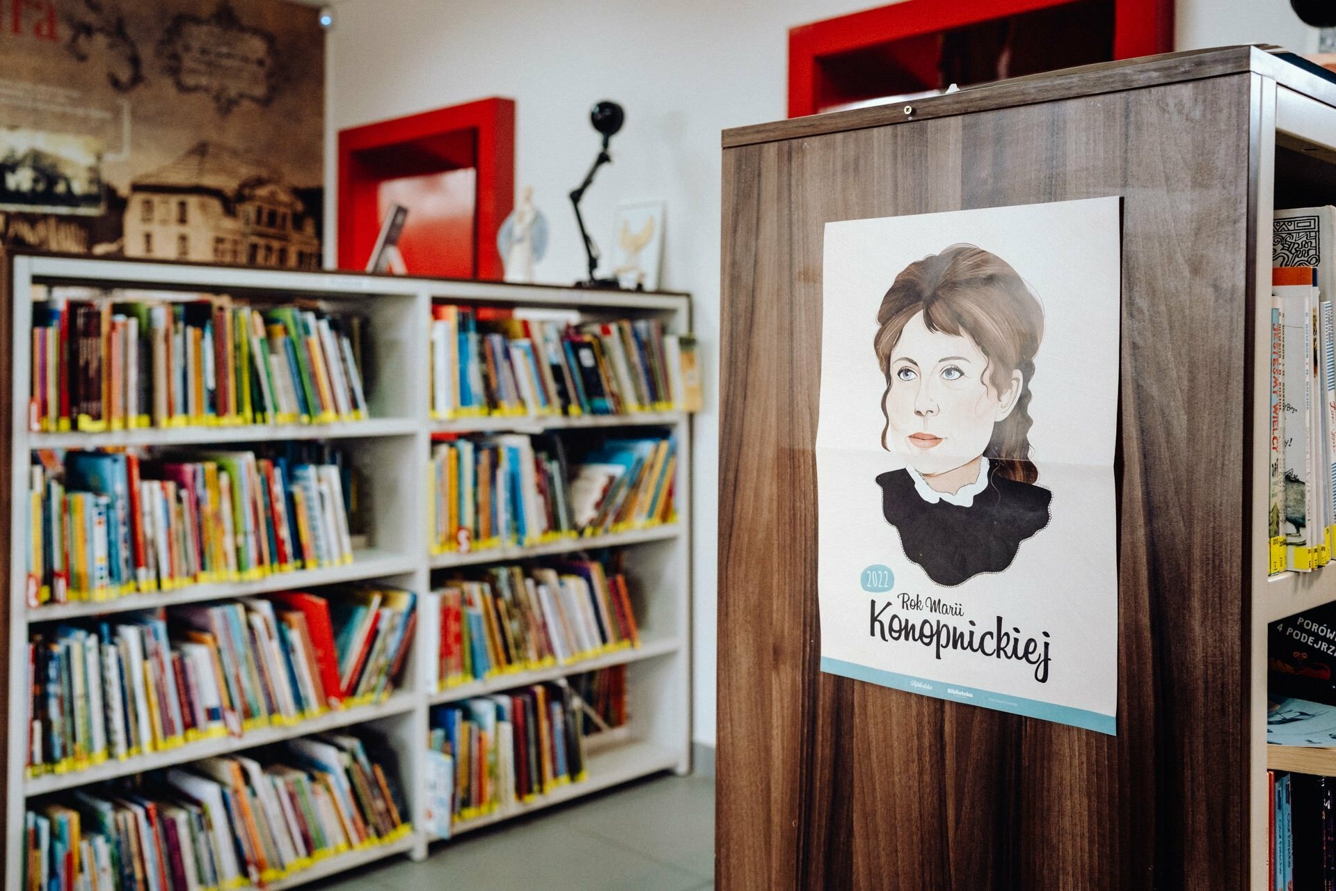 The interior of the library with shelves filled with books. We focus on a wooden cabinet on which there is a poster depicting a woman with brown hair and a serious expression. The caption reads, "Kob Maria Konopnicka." The red doorframes and various decorations provide the perfect backdrop for any event photographer Warsaw.   