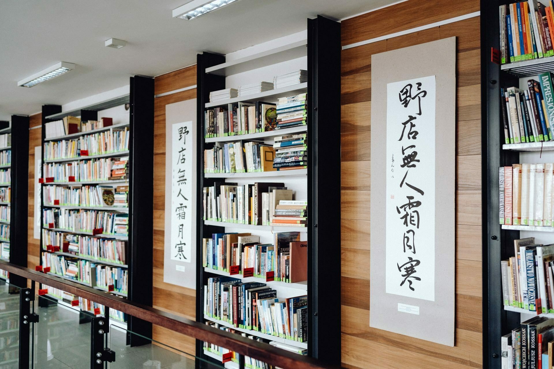 Modern library with high shelves filled with books and colorful covers. The shelves are integrated into the wooden wall with vertical panels in a light color. Between the shelves hang large works of traditional Asian calligraphy art, perfect for an event photographer Warsaw capturing photo coverage of an event.  