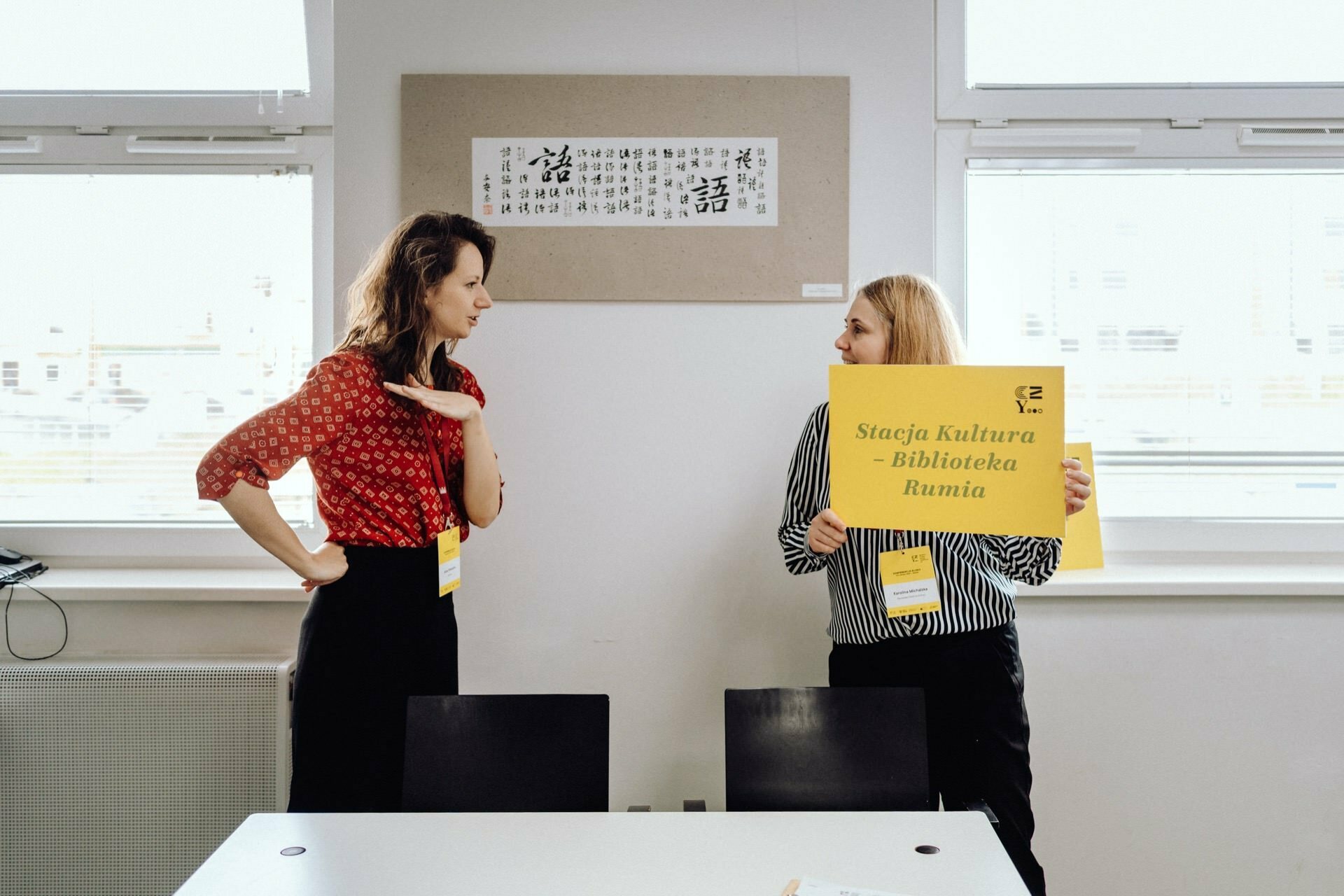 Two women stand in a room next to a table. The woman on the left, wearing a red shirt, gestures with her hand. The woman on the right, wearing a black and white striped shirt, is holding a yellow sign that reads "Culture Station - Rumia Library." This photo report from the event perfectly captures their engaging conversation.   