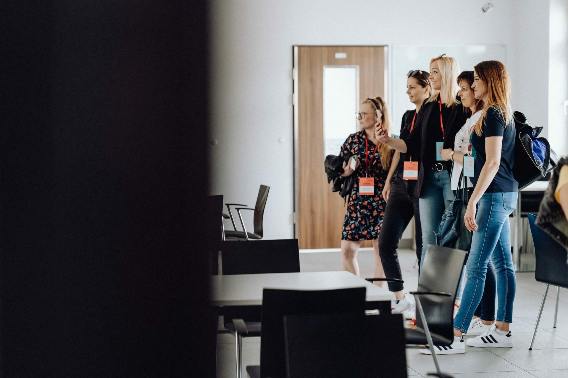 Four women stand and pose for a photo in a room with tables and chairs. They are wearing casual clothes and conference badges. The room has a modern, minimalist decor with wooden doors and white walls in the background - a perfect place that any event photographer Warsaw will capture.  