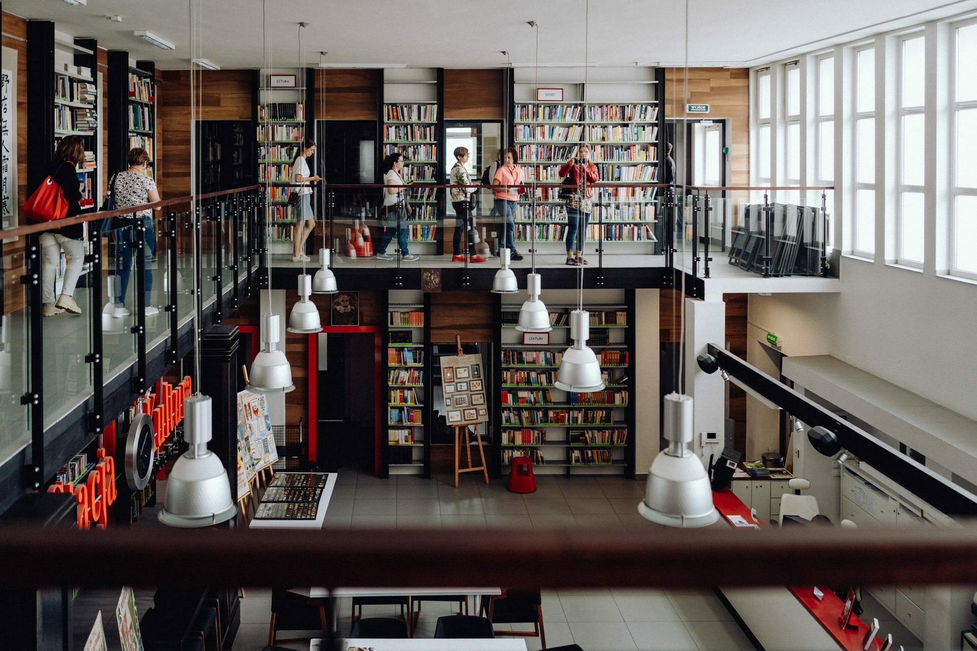 A spacious, modern library with a two-story layout. The lower floor has bookshelves and work areas, while the upper floor has a glass balustrade and additional bookshelves. Several people are browsing and reading books, perhaps preparing for a photo essay of the event. Large windows allow natural light to fill the space.   