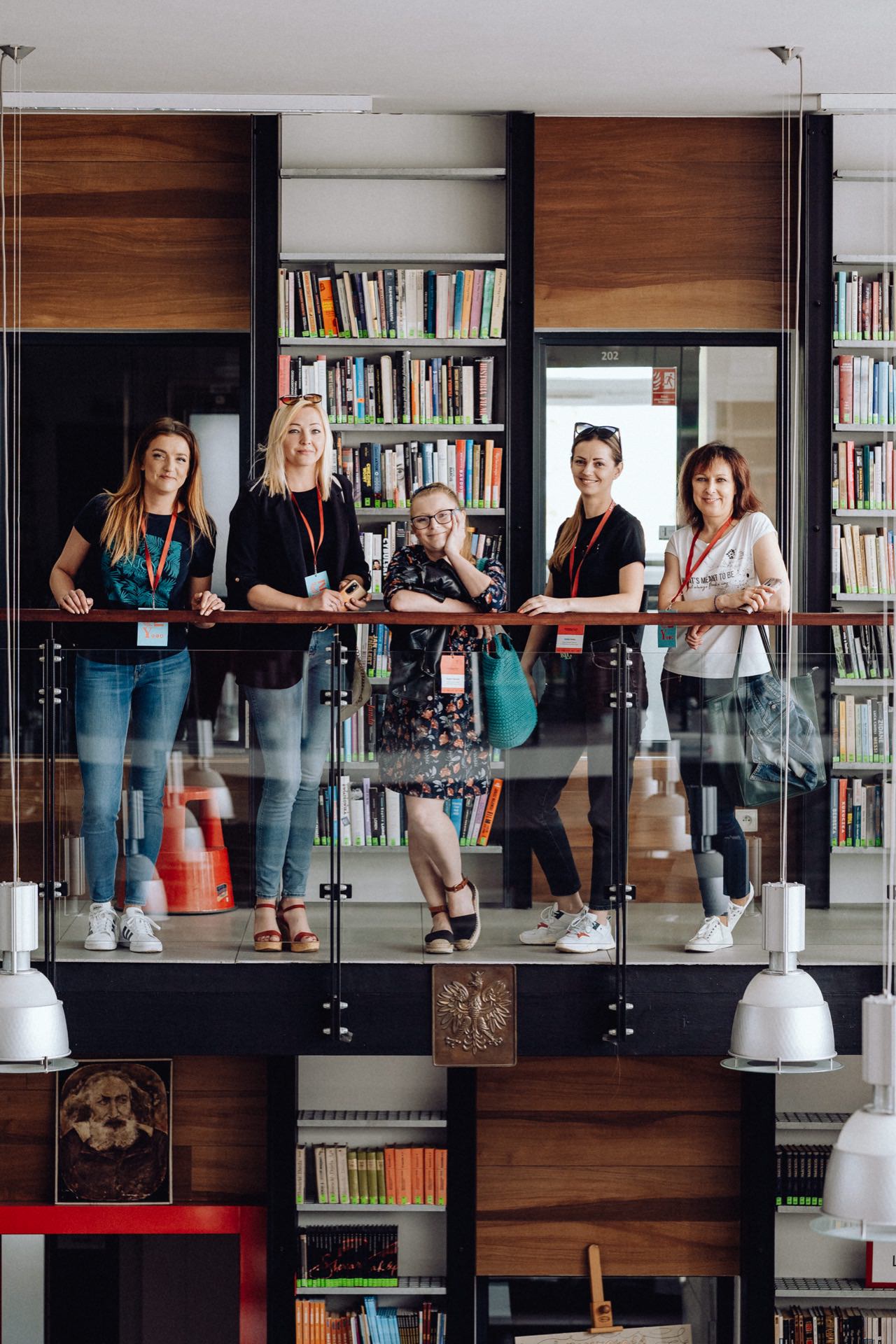 Five people stand on the floor of a modern library, leaning against a glass barrier. Below them are stylish bookshelves that hold a variety of books. The group gives the impression of being relaxed, each wearing a lanyard, suggesting a professional or academic setting - perfect for an event photo shoot in Warsaw (event photographer Warsaw).  