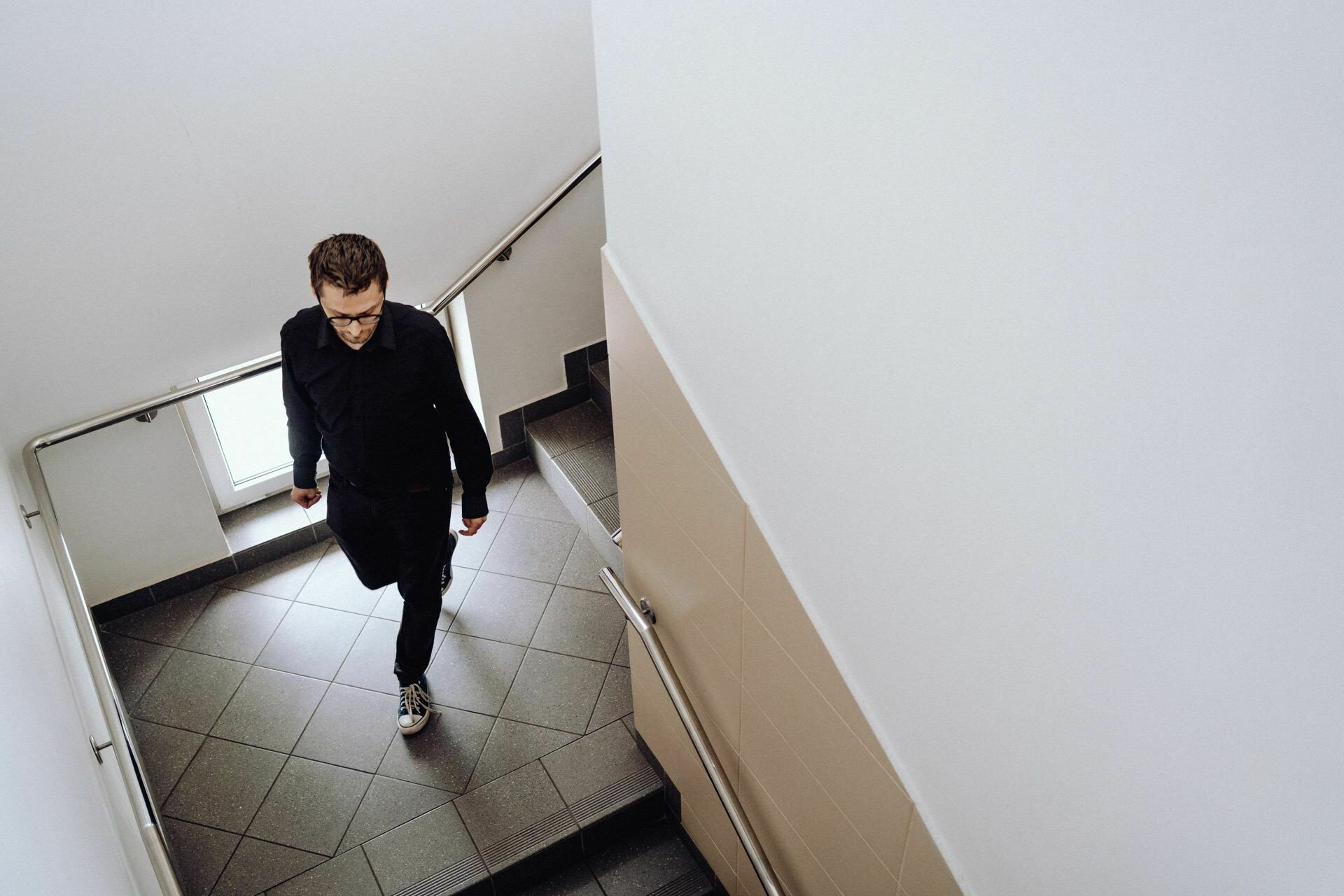 A man wearing glasses, dressed in a black shirt and dark pants, walks up a tiled staircase in a minimalist room. He looks down, and the scene is lit by natural light - perfectly captured by an event photographer from Warsaw. 