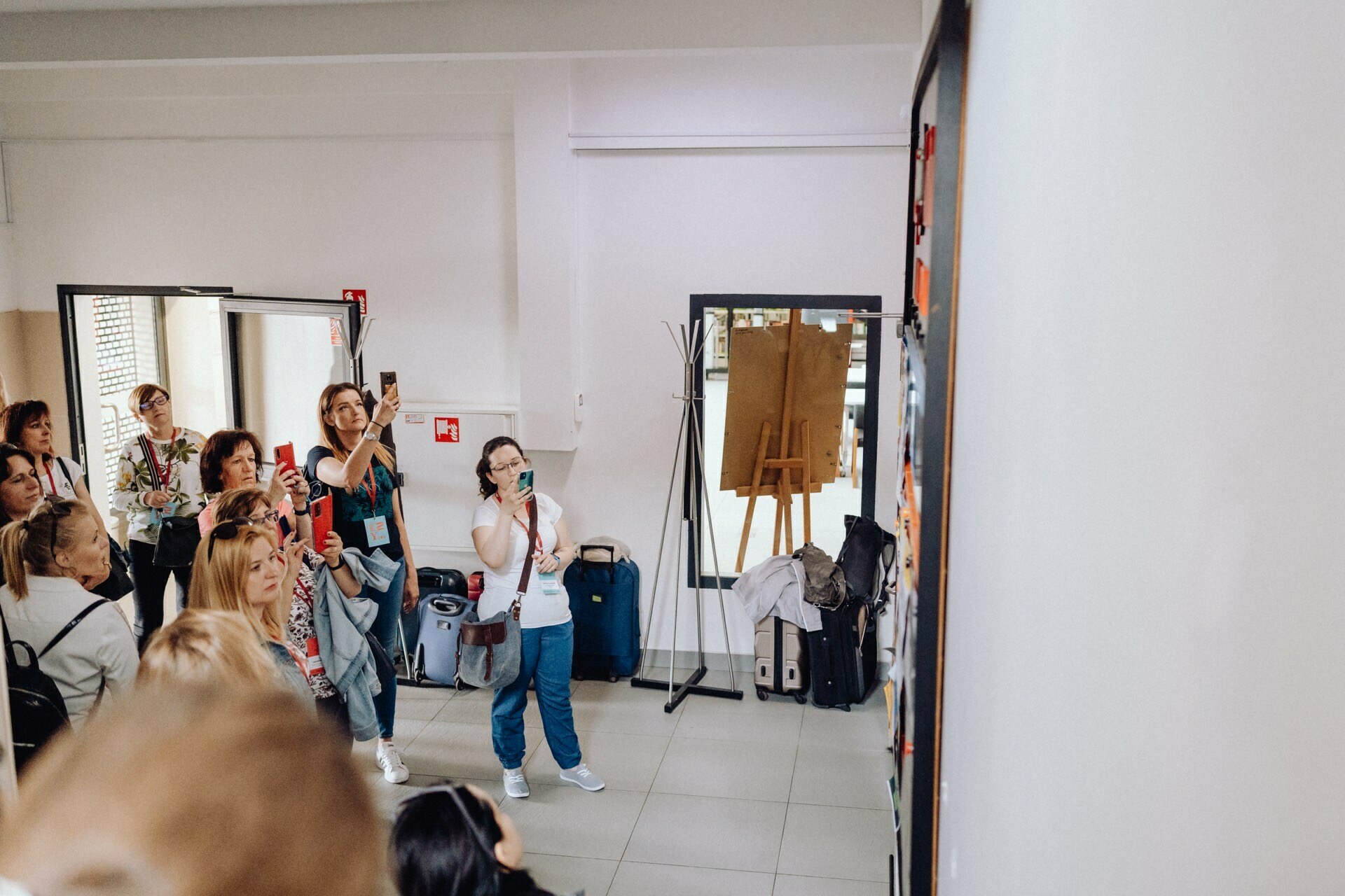 A group of people are standing in a room with white walls and looking intently at something to the right, out of sight. A few are taking photos or videos with their phones, capturing what appears to be a photo essay of the event. An easel and several bags can be seen in the background, as well as open doors and windows.  