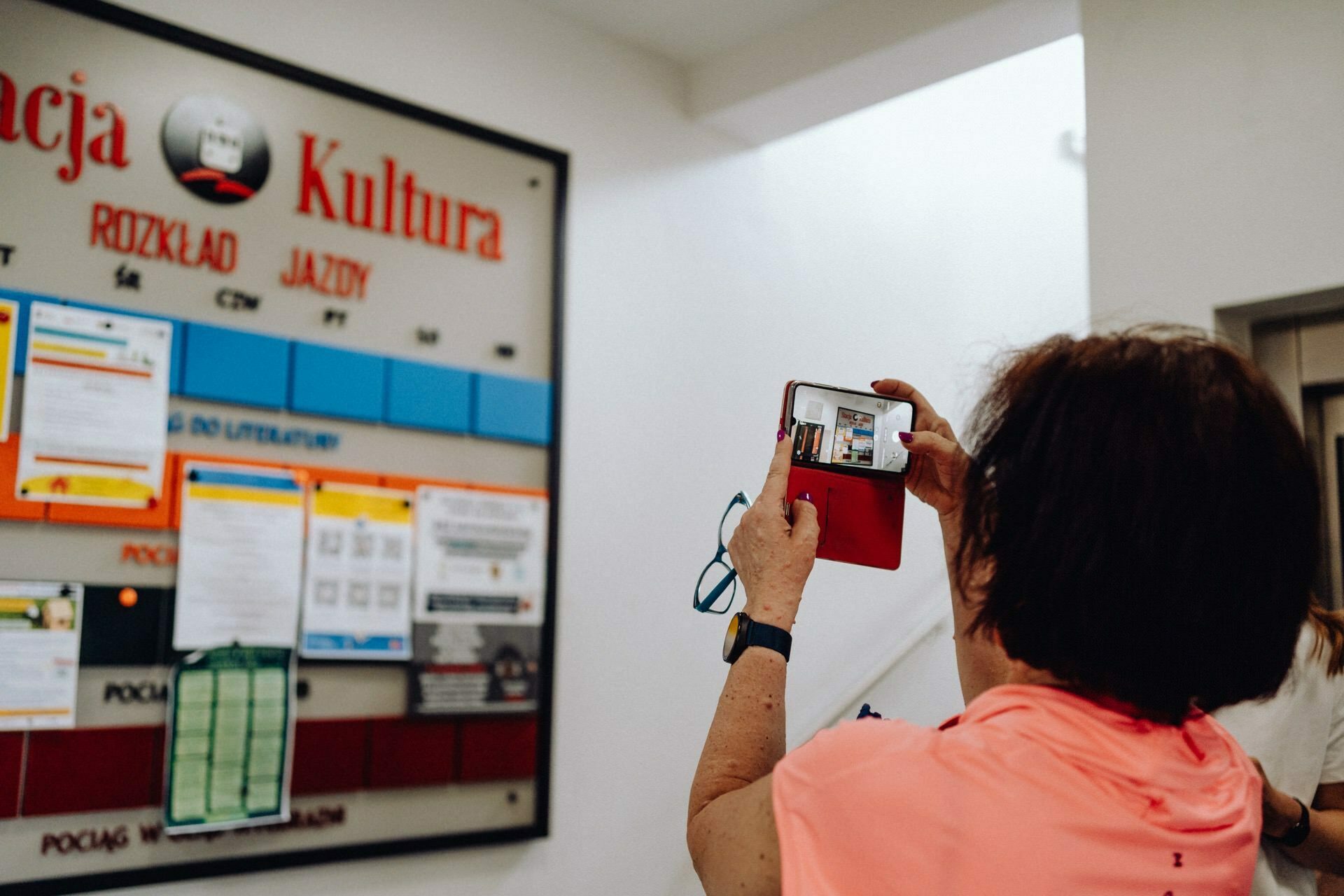 A person wearing a pink shirt and wristwatch is taking a photo with a smartphone of a large bulletin board, perhaps for a photo essay on the event. The board shows various schedules and notifications, and the action takes place in a room, probably a train station or other public place. 