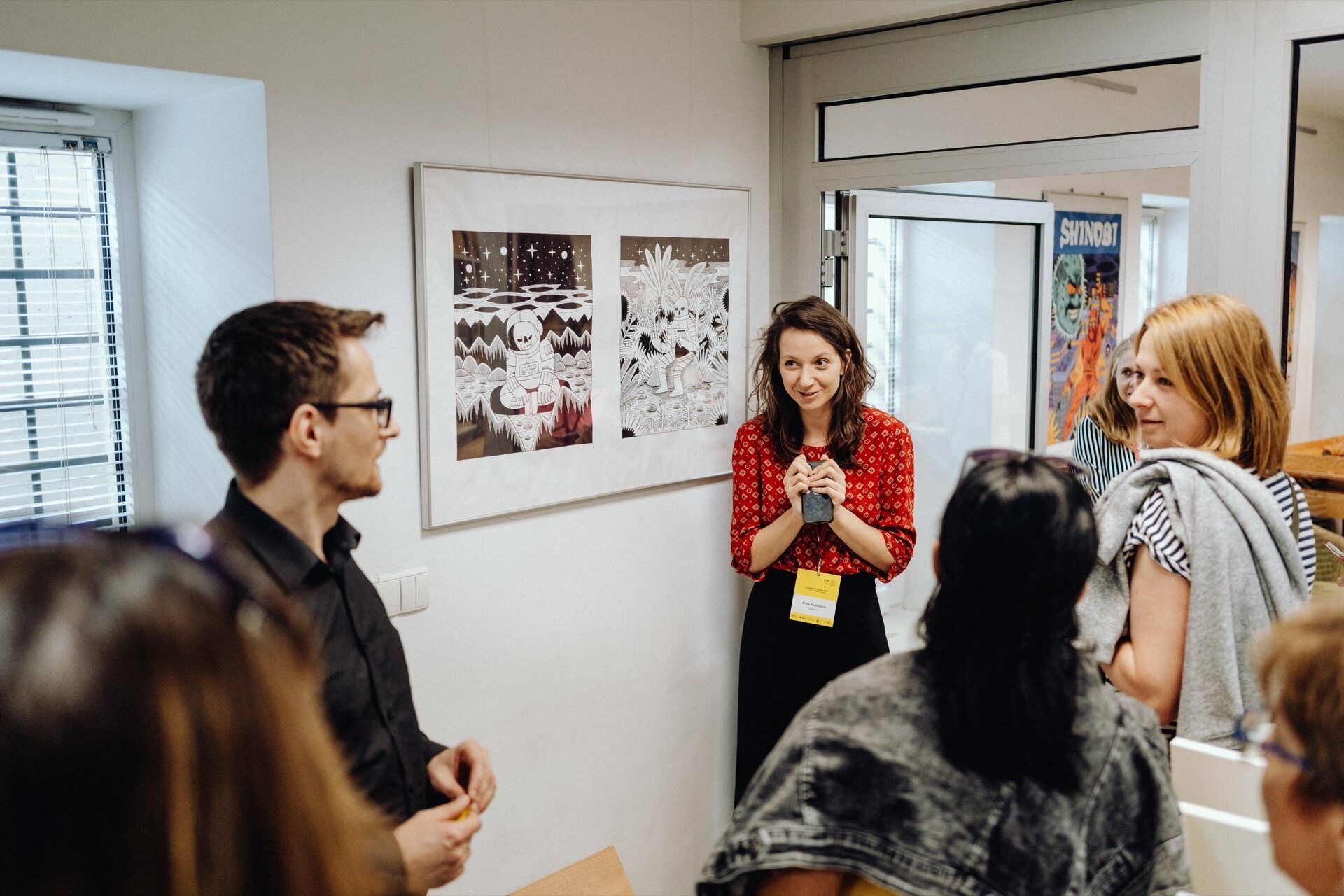 A woman in a red shirt stands and talks to a small group of people in an art gallery. Behind her hangs a framed print. Other attendees listen intently, including a man in a black shirt and several women, one in a striped shirt. The photographer for the event captures the moment perfectly.   