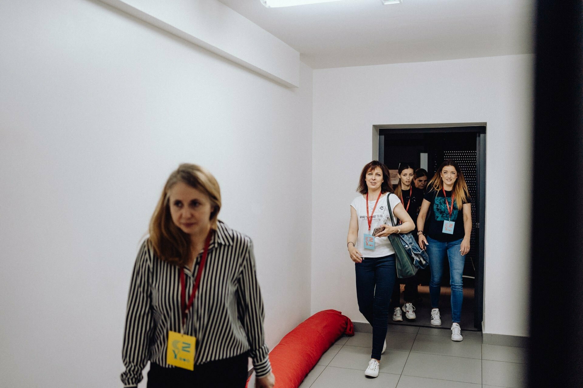 A group of people walking around the room, led by a woman in a striped shirt with a yellow name badge. Behind her are three women with event badges, one carrying a blue bag. The corridor is well-lit, with white walls and a red padded object on the floor, perfect for a photographer at the event to take pictures of the event.  