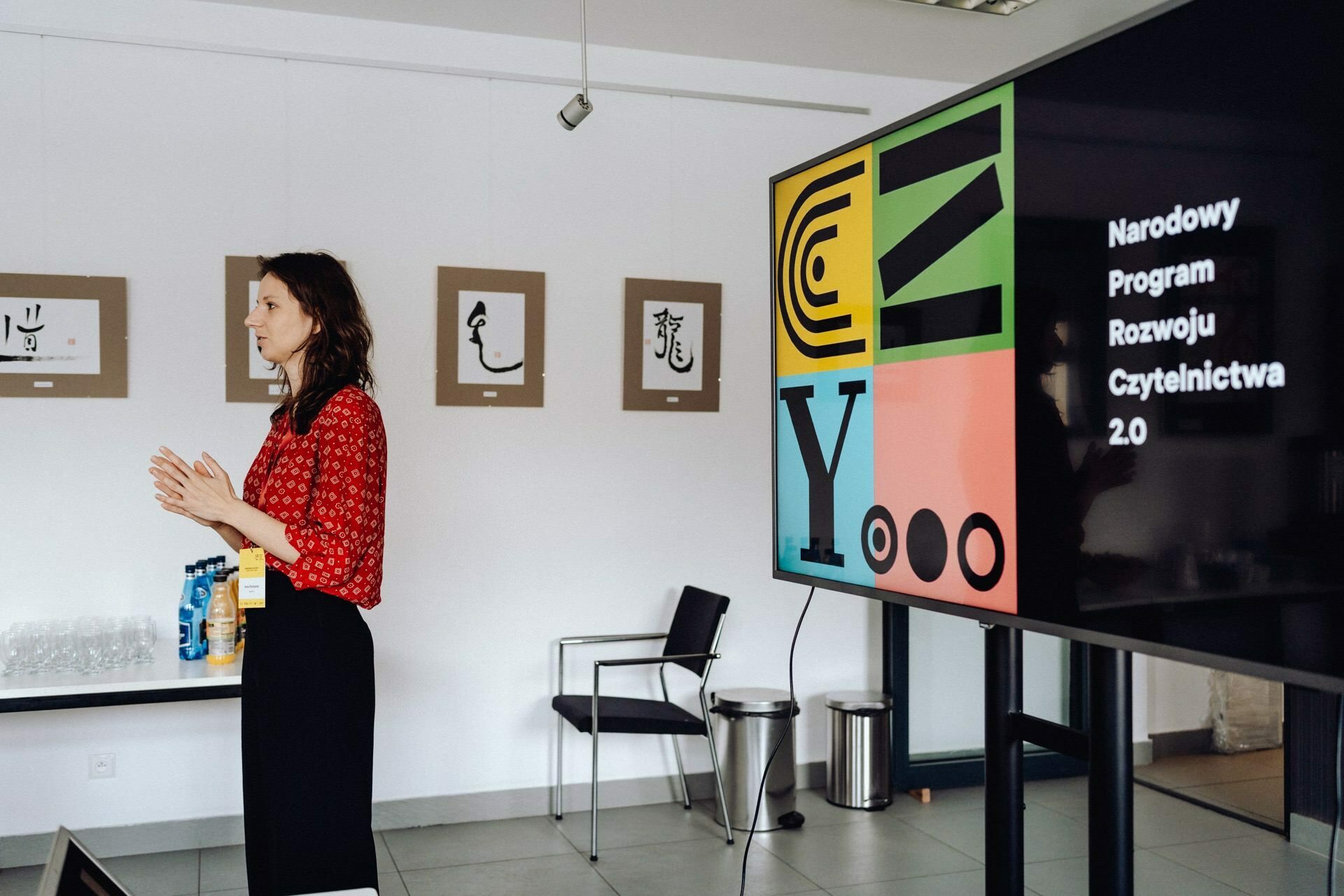 A woman in a red patterned blouse stands in a room and gives a presentation. To her left is a screen with the words "National Reading Development Program 2.0." On the wall behind her you can see framed works of art, and next to her several empty chairs and a table, perfectly captured by a photographer from Warsaw.  