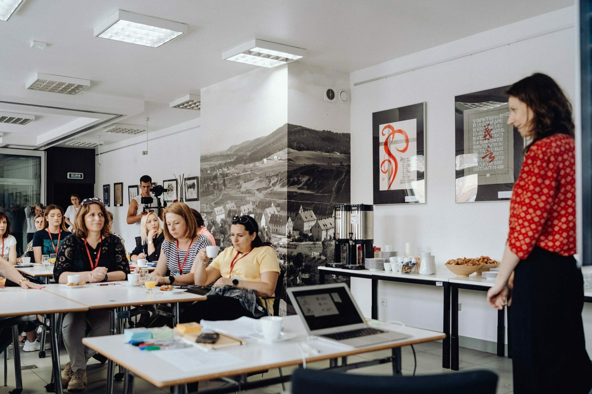 A woman in a red blouse stands at the front of the classroom and addresses a group of people seated at tables. The room has modern lighting, artwork on the walls and snacks on a side table. A projector with a laptop is set up to assist in the presentation - a stage perfect for any event photographer Warsaw.  