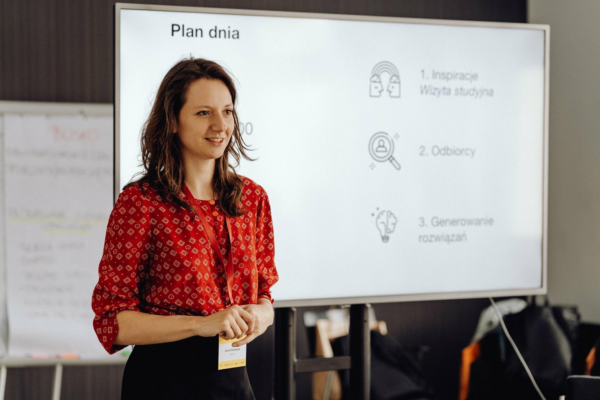 A woman in a red patterned blouse is standing in front of a presentation screen that displays the daily schedule. On the screen are three dots with icons: "Study Visit Inspiration," "Recipients" and "Network Generation." A flipchart captured by a photographer at the event is visible in the background.  