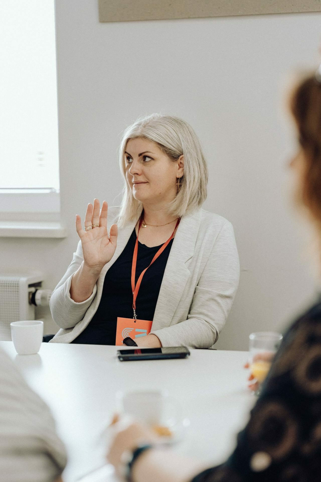 A woman with shoulder-length gray hair, wearing a white jacket and red lanyard, sits at a table and raises her hand. She has a neutral expression on her face, with two white cups and a smartphone in front of her. In the foreground are two blurry figures, capturing the essence of the event photo report by event photographer Warsaw.  