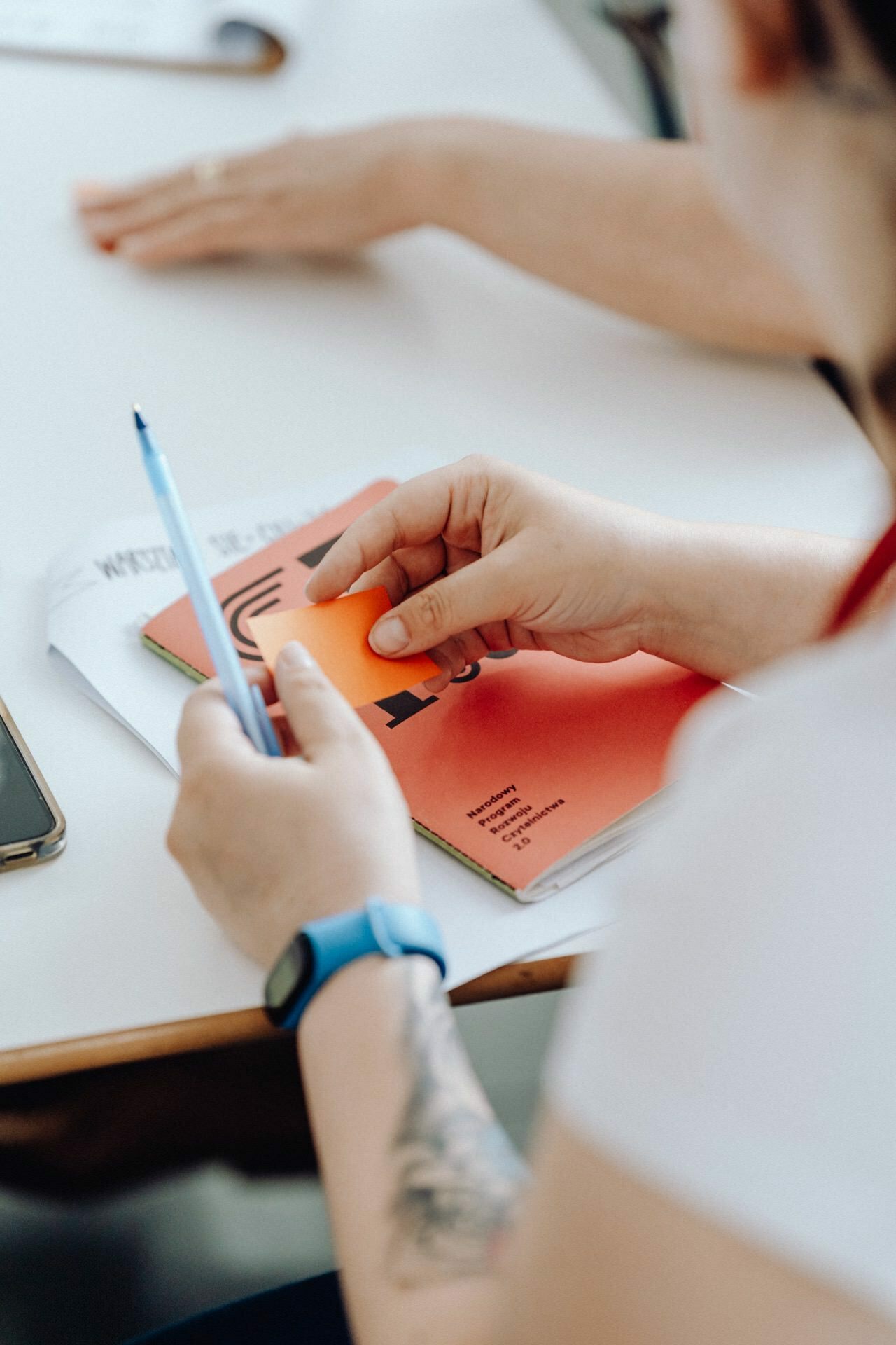 A person wearing a white shirt and a blue watch, holding a blue pen and an orange piece of paper, with a tattoo visible on his forearm. The person is sitting at a table with papers and a phone. Another person's hands are visible in the background, part of a dynamic photo story of the event captured by an experienced event photographer warsaw.  