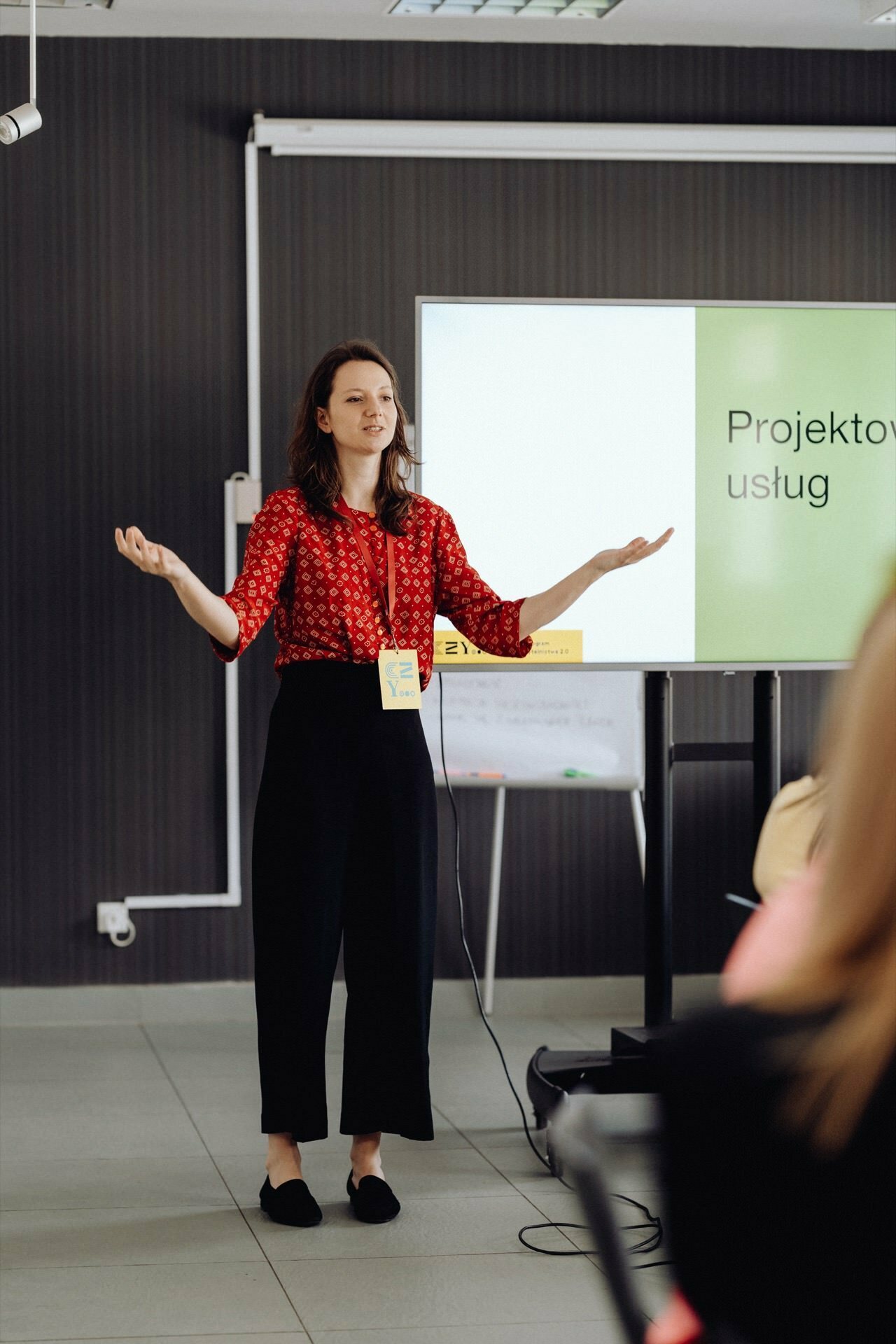 A woman in a red patterned blouse and black pants is standing in front of a presentation screen that reads "Projektov services." She speaks with open arms, addressing the audience. A lanyard hangs around her neck, perfectly captured by a photographer at the event during this photo essay of the event.  