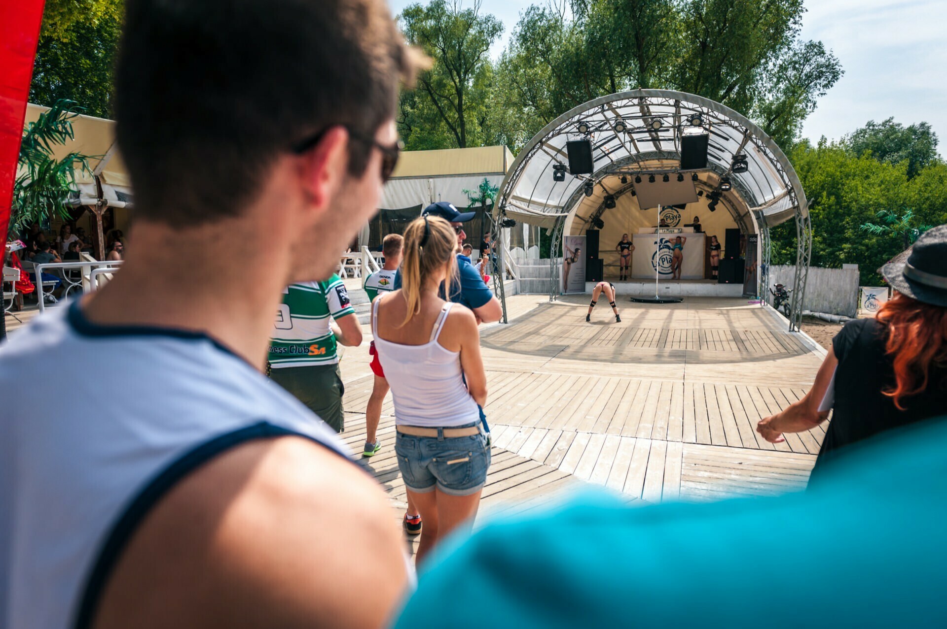 People gather at an outdoor event, watching a performance on a small stage with a metal arch. In the background are trees and buildings, and in the foreground are various figures in casual summer attire, perfectly capturing the atmosphere of the Photo Fair. 