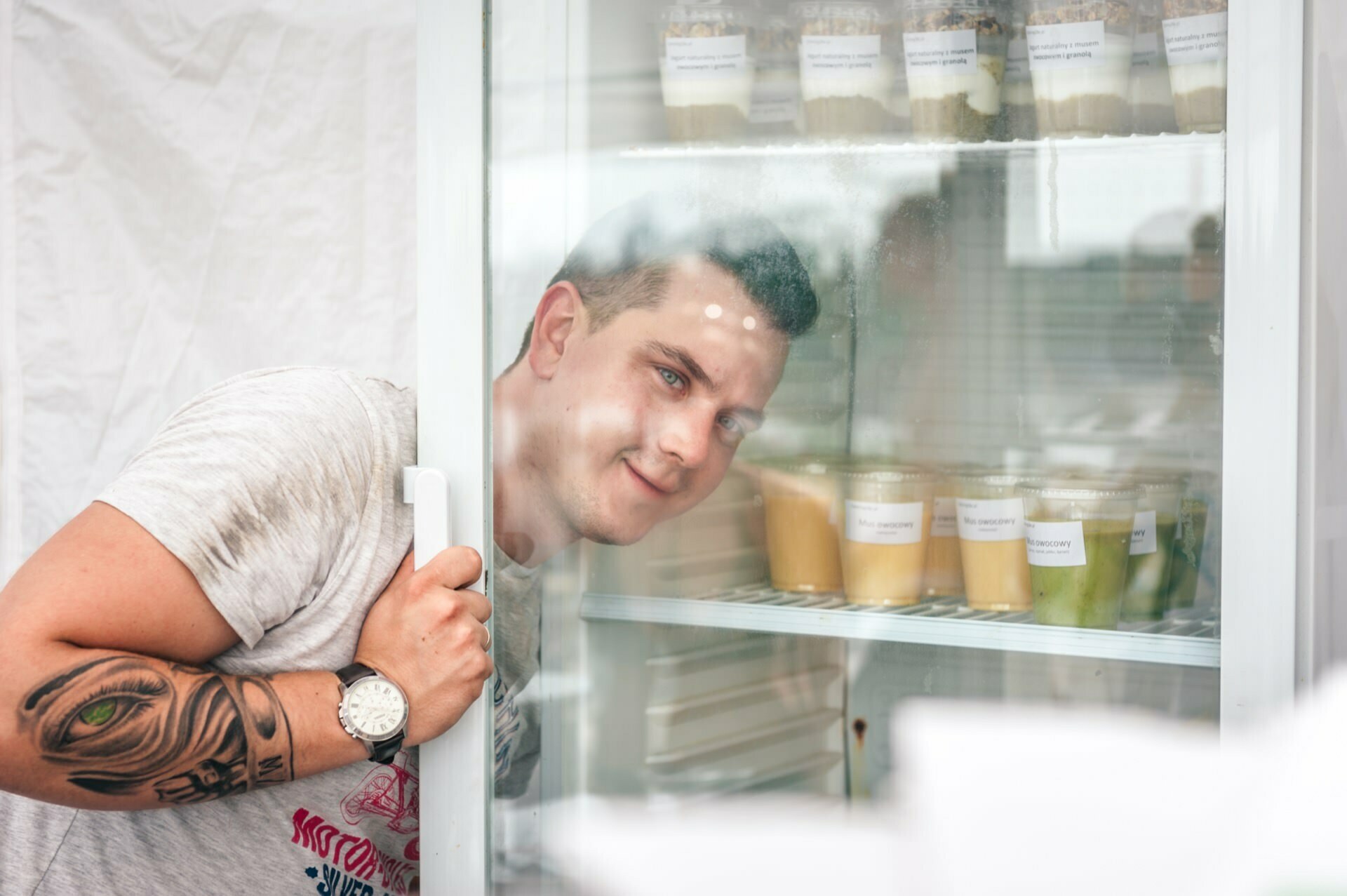 A man with an arm tattoo and a watch smiles as he looks into a refrigerator filled with various food containers, some with visible labels. He holds the refrigerator door slightly ajar. The scenery seems casual and full of light, like a photo essay from a trade show capturing everyday moments.  