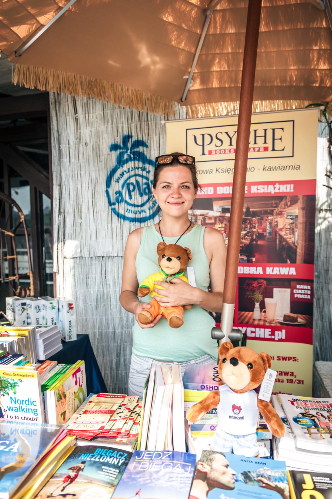 A smiling woman stands at a book stand under an umbrella. She is holding a teddy bear and is surrounded by colorful books. A sign behind her with the words "PSYCHE" and an advertisement for a bookstore and cafe captures the essence of the photo fair. The booth features various books and another teddy bear.   