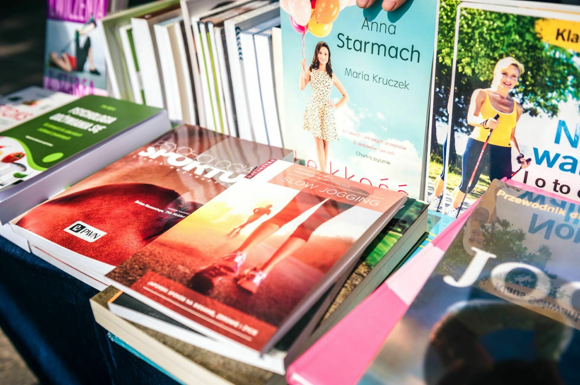 A display table with a variety of books for sale, showcasing a vibrant photo spread from the fair. Well-known titles include a book titled "Anna Starmach," a book featuring a running person, and other books related to health and fitness. The carefully arranged books are colorful and inviting.  