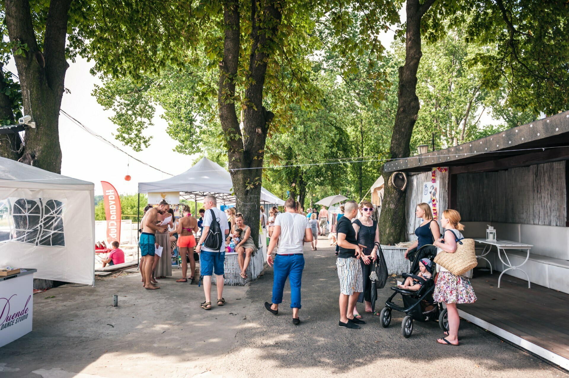 Photo report of a fair of people gathered outdoors at a small market or festival, with various booths and tents. Some browse through items, others talk or walk around. Shade trees, and to the right, mothers  