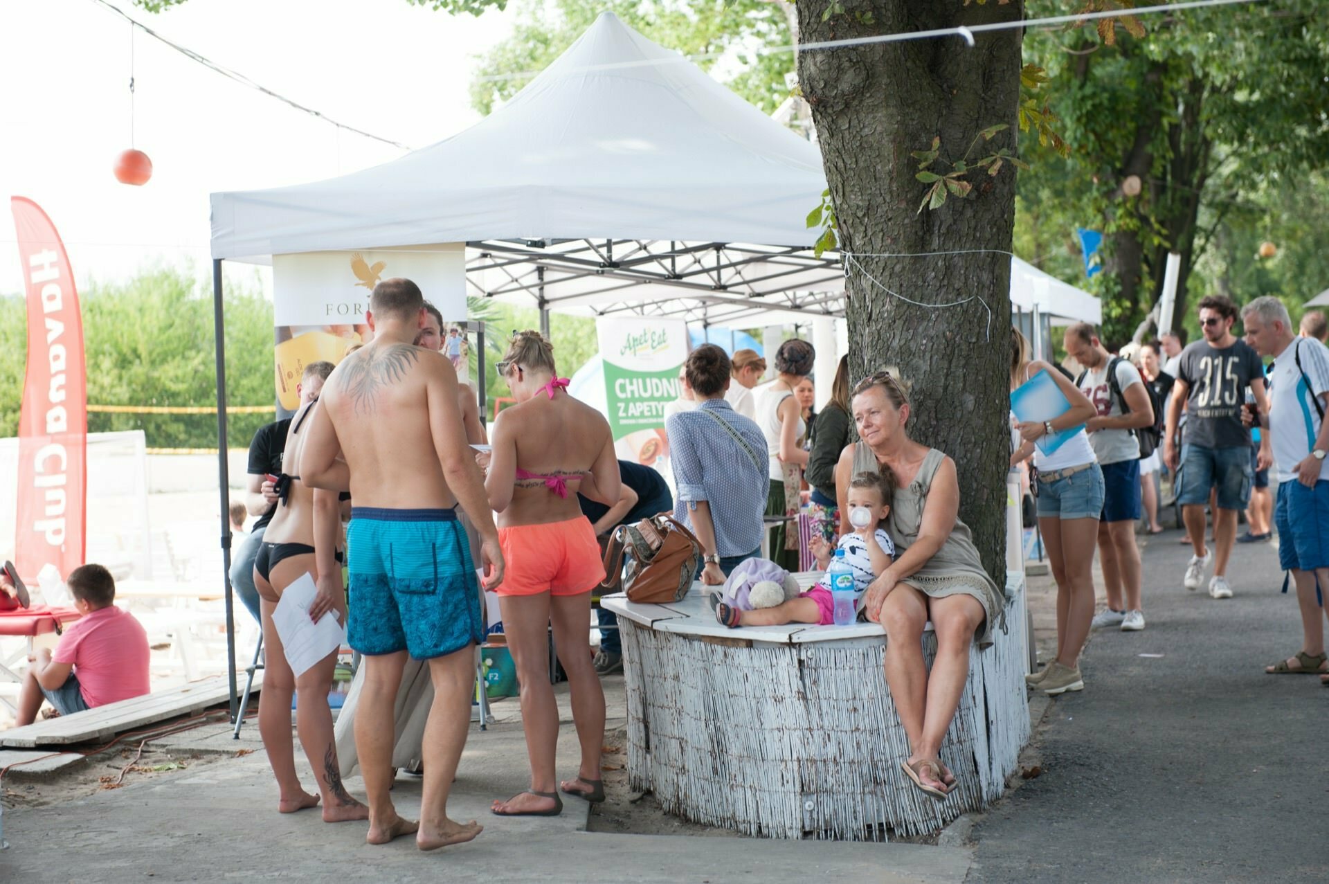 A group of people have gathered around a white tent at an outdoor event near a large tree. Some are talking, others are looking through documents, capturing the essence of the photo fair. A woman holding a baby sits on a white barrel, and people are dressed in summer clothes, suggesting a warm day.  