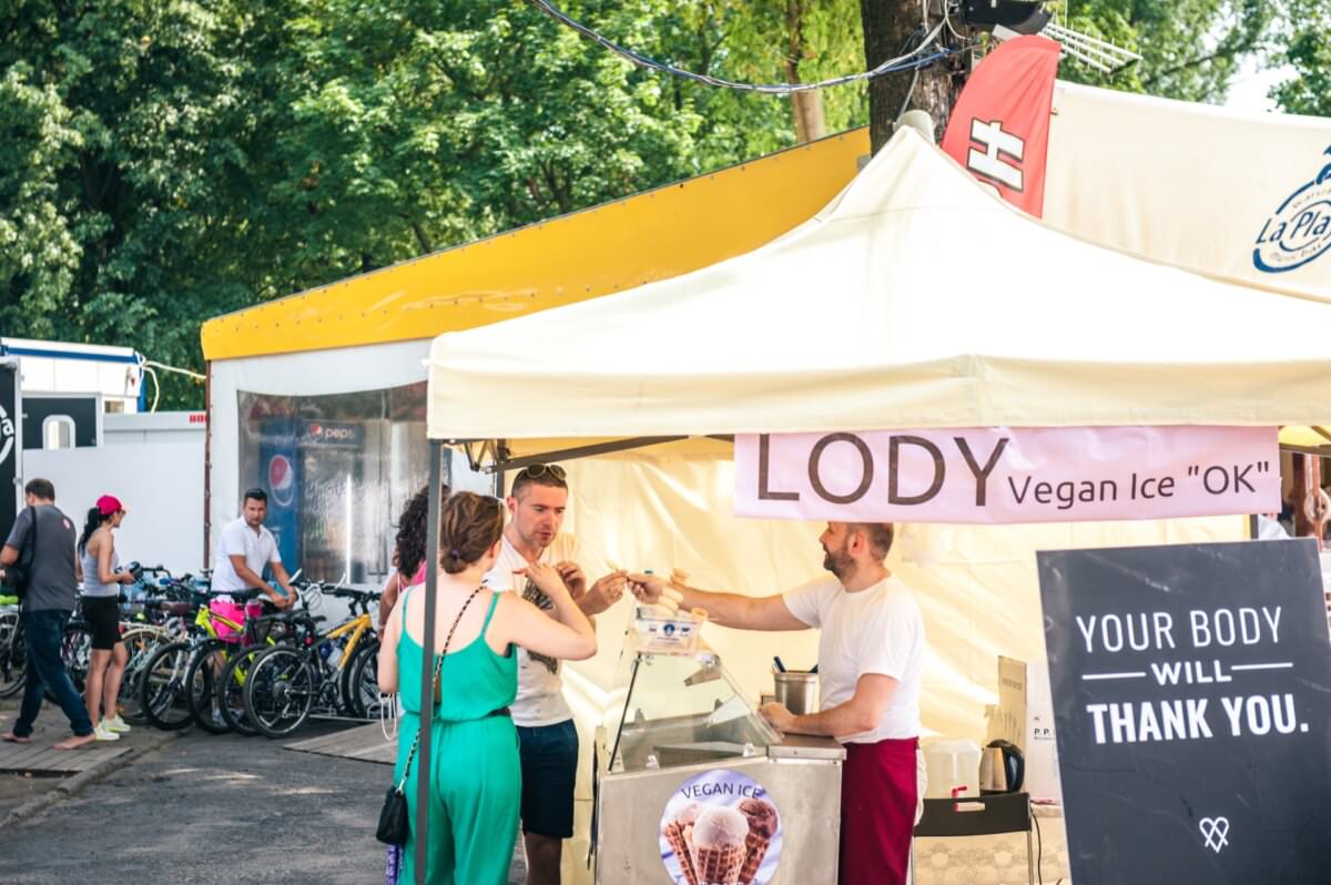 Słoneczny targ na świeżym powietrzu z ludźmi zgromadzonymi wokół jasnożółtego namiotu z napisem „LODY Vegan Ice „OK”. Sprzedawca w namiocie serwuje klientom wegańskie lody. W tle zaparkowane są rowery, a drzewa dają cień. To idealny dzień na fotorelację z targów!