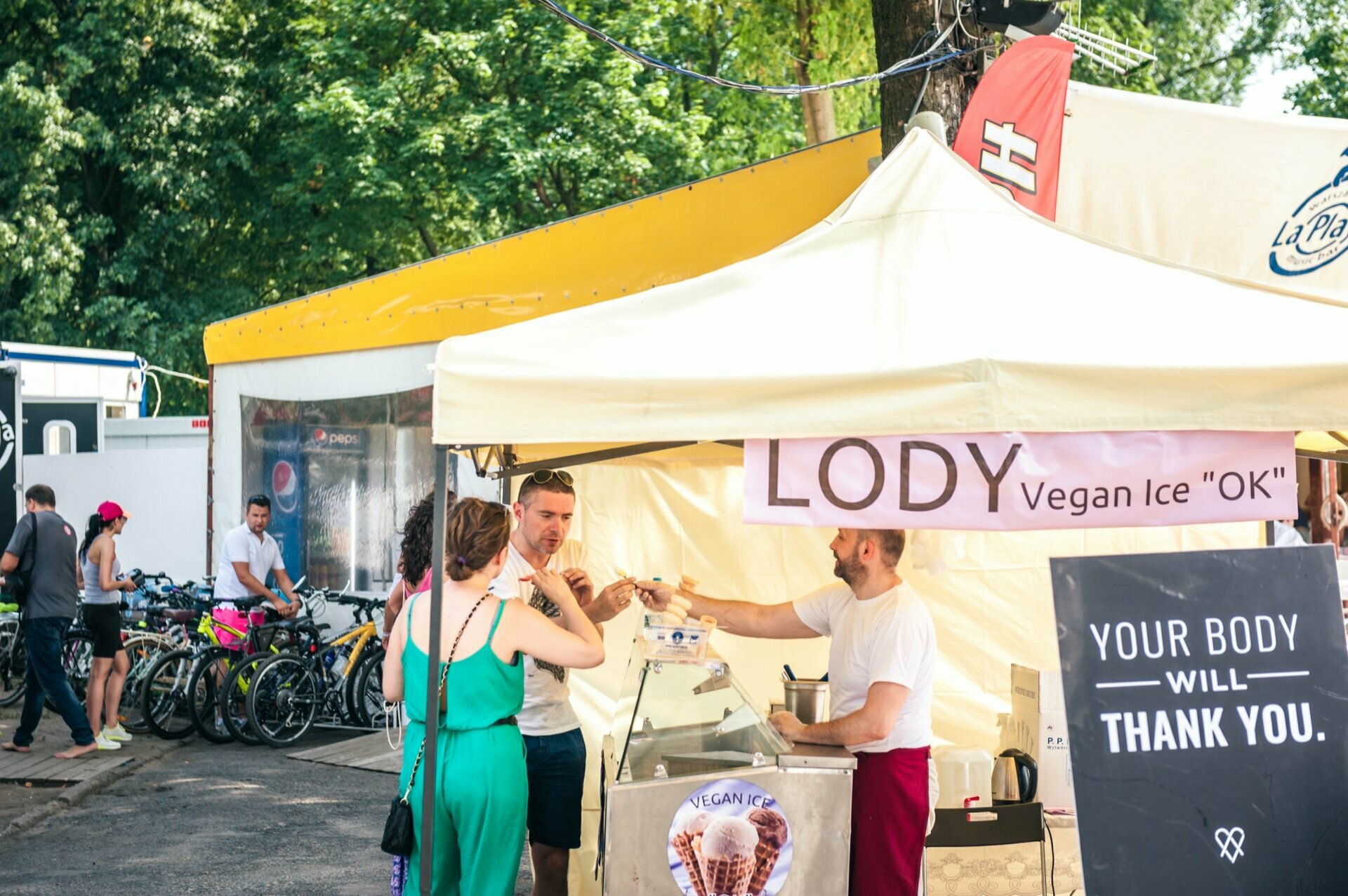 A sunny outdoor market with people gathered around a bright yellow tent with a sign that reads "Vegan Ice Cream "OK". A vendor in the tent serves vegan ice cream to customers. Bicycles are parked in the background and trees provide shade. It's a perfect day for a photo report of the fair!   