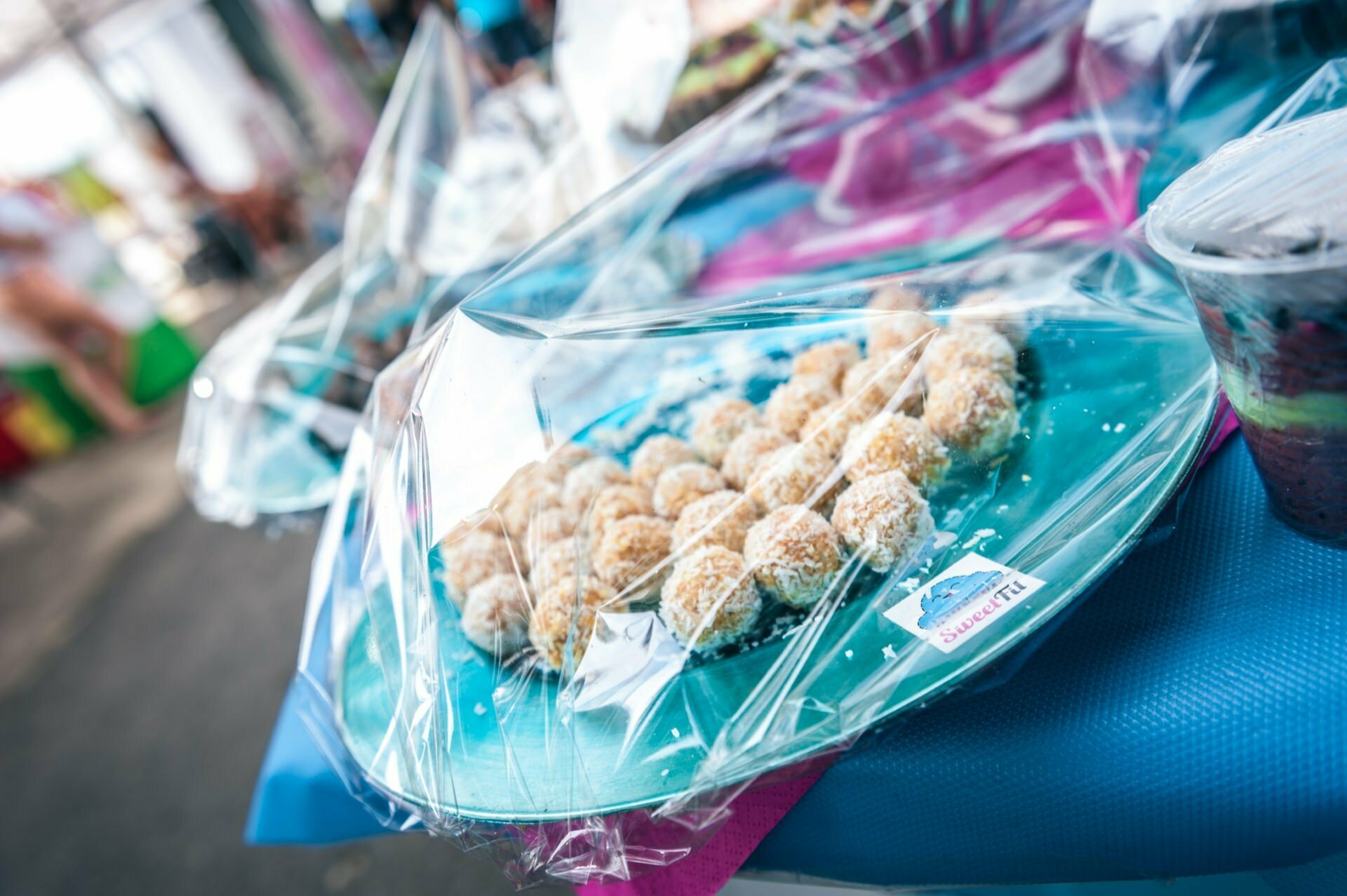 Close-up of a tray filled with coconut-covered, foil-wrapped round treats. The tray stands on a table with colorful covers. In the background, blurry images of more foodstuffs and people, suggesting an outdoor market or fair setting - perfect for a photo report of a fair.  