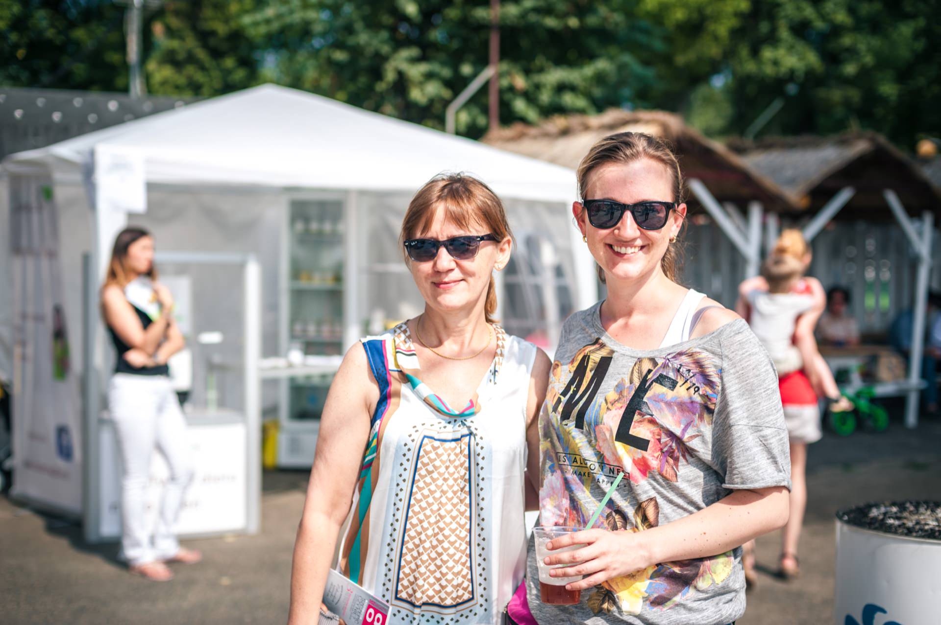 Two women are standing outside in casual summer attire, both wearing sunglasses and smiling at the camera. The woman on the left is wearing a patterned sleeveless top, the woman on the right is wearing a graphic T-shirt and holding a plastic cup. In the background you can see booths and trees from the photo fair.  