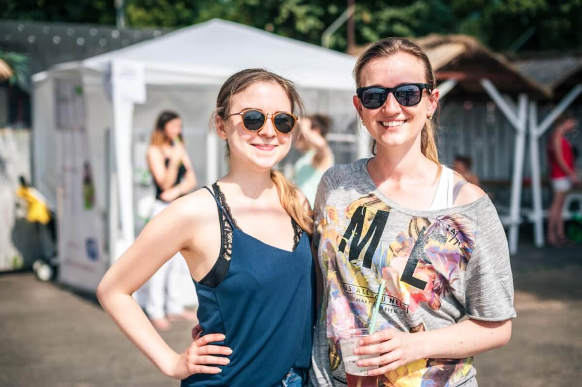 Dwie kobiety uśmiechają się i pozują na zewnątrz w słoneczny dzień. Oboje noszą okulary przeciwsłoneczne; kobieta po lewej ma granatowy top bez rękawów, kobieta po prawej ma na sobie szary top z kolorowym tekstem. W tle widać biały namiot i innych ludzi, co uchwyciło tętniącą życiem fotorelację z targów.