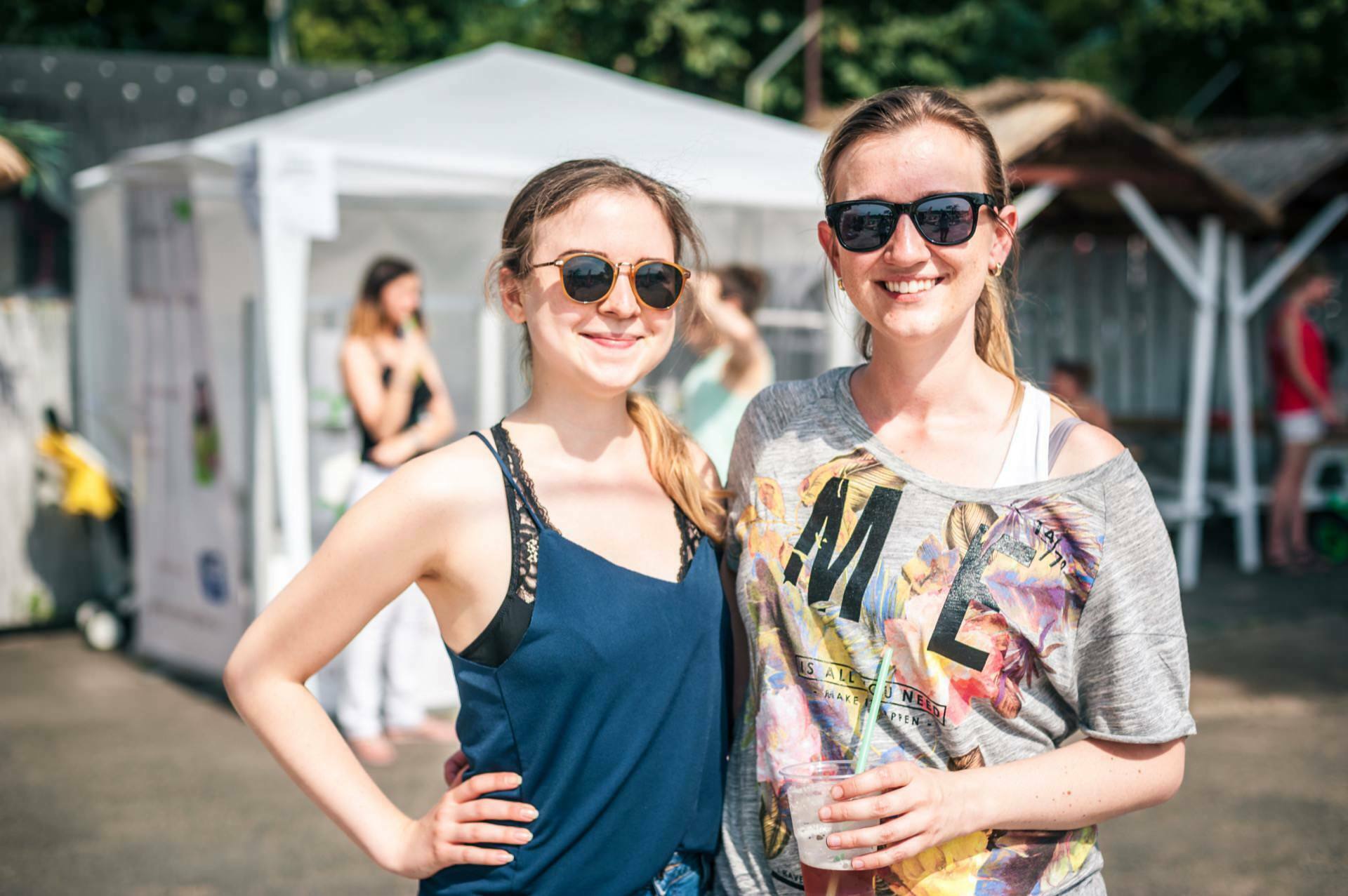 Two women are smiling and posing outside on a sunny day. Both are wearing sunglasses; the woman on the left is wearing a navy blue sleeveless top, the woman on the right is wearing a gray top with colorful text. A white tent and other people can be seen in the background, capturing a vibrant photo-op of the fair.  