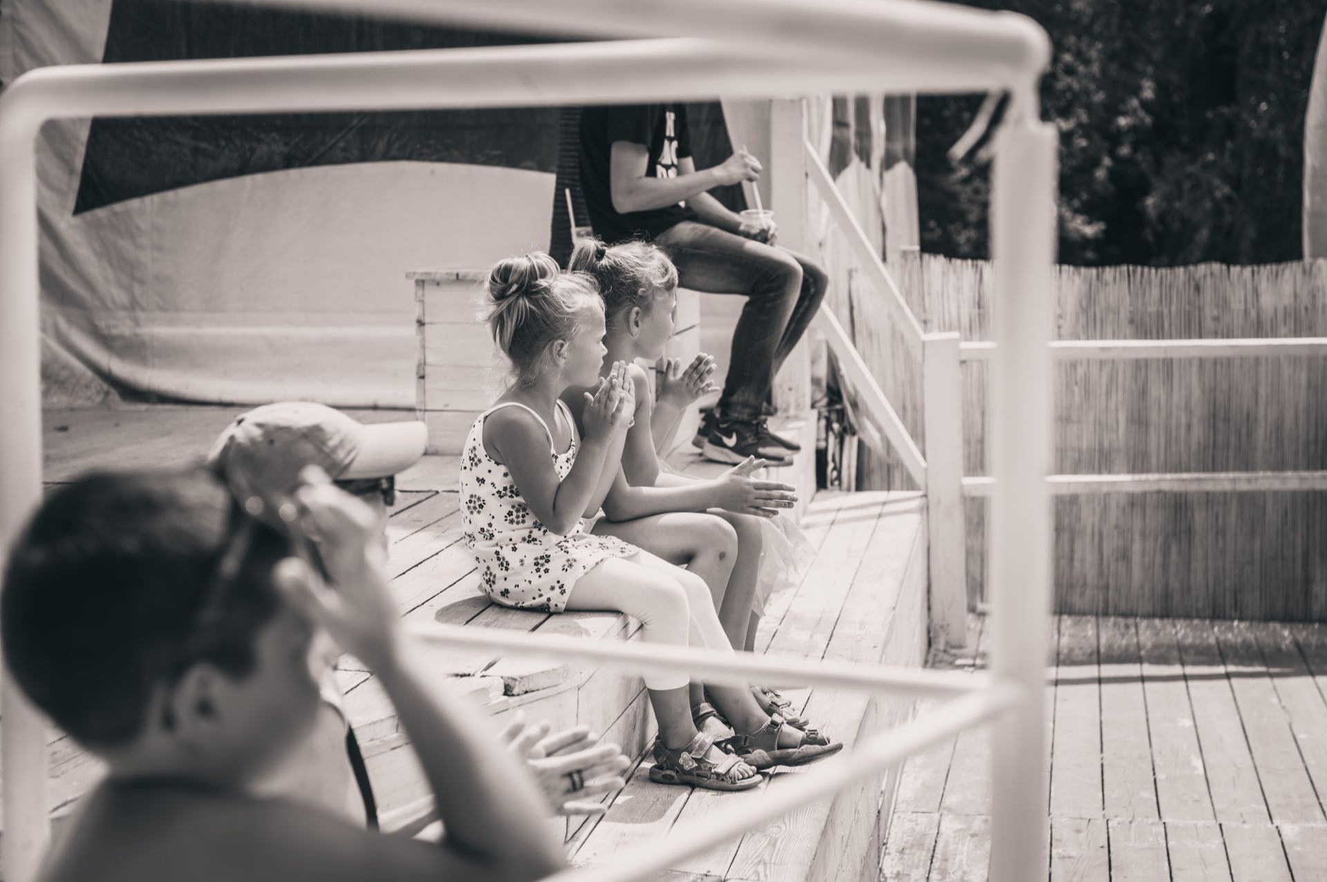 Black and white photo of three children sitting on a wooden terrace, clapping and watching something out of frame. An adult stands nearby, leaning against the railing. The terrace is surrounded by a wooden fence and partially shaded. This quiet moment evokes the warmth of a photo fair, but everyone seems to be outside.   