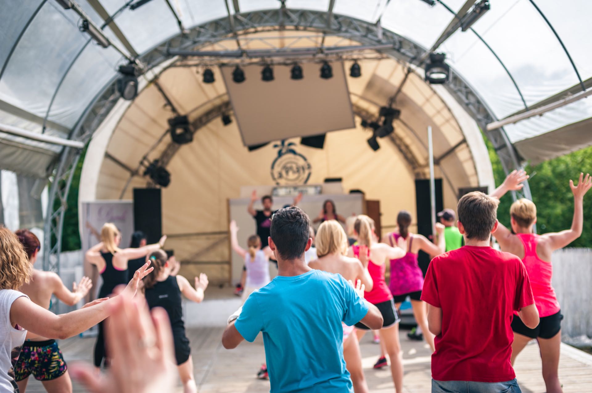 A group of people participate in an outdoor fitness class under the large canopy of a tent. They follow the instructor's movements while wearing casual workout clothes. The surroundings are bright and vibrant, with a stage and various fitness equipment in the background, perfect for photo opportunities at the fair.  