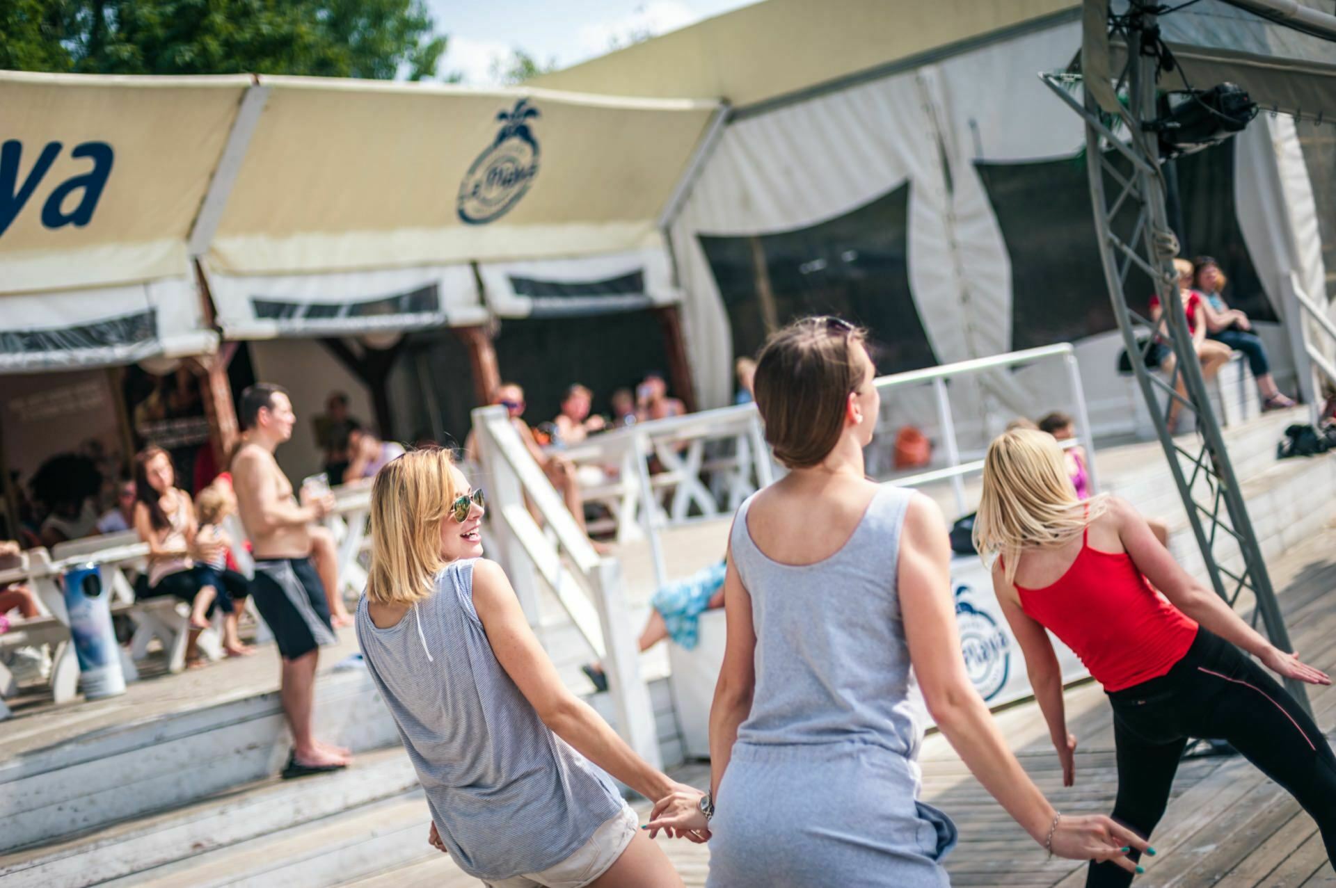 Three women, two in gray and one in red, dance outdoors on a wooden terrace. Behind them, people sit under a pavilion with white tents and signs. Others stand nearby, enjoying the sunny weather and various activities - a vibrant scene perfect for a photo essay of the fair.  