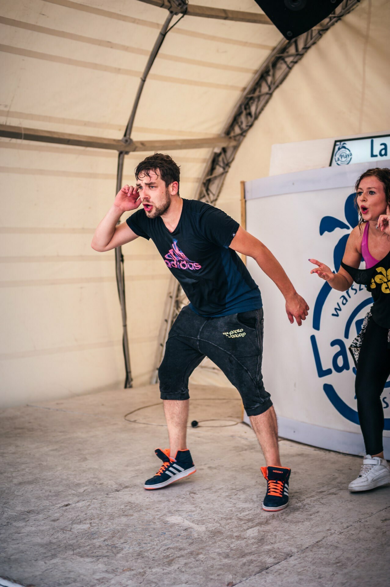 The photo report from the fair shows a man and a woman energetically performing on stage inside a large tent. The man is wearing a black T-shirt, black shorts and black sneakers with orange accents, and poses as if attentively listening. The woman next to him appears to be engaged in a dynamic performance.  