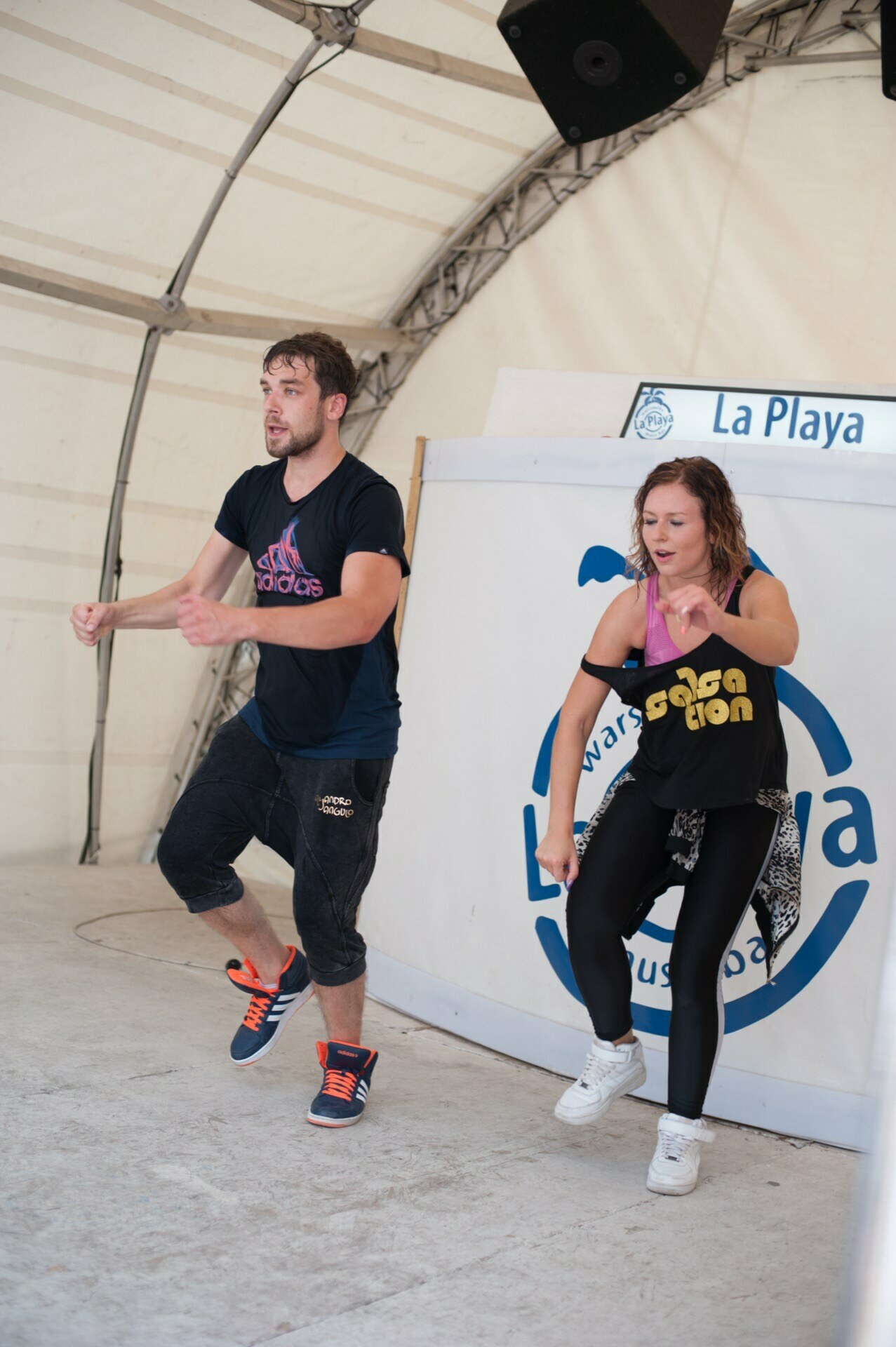 Two people are dancing in a room with a white tent canopy. The man on the left is wearing a navy blue t-shirt and shorts, the woman on the right is wearing a black T-shirt and leggings. Both appear to be moving in mid-dance, with energetic expressions capturing the lively spirit of the photo fair.  