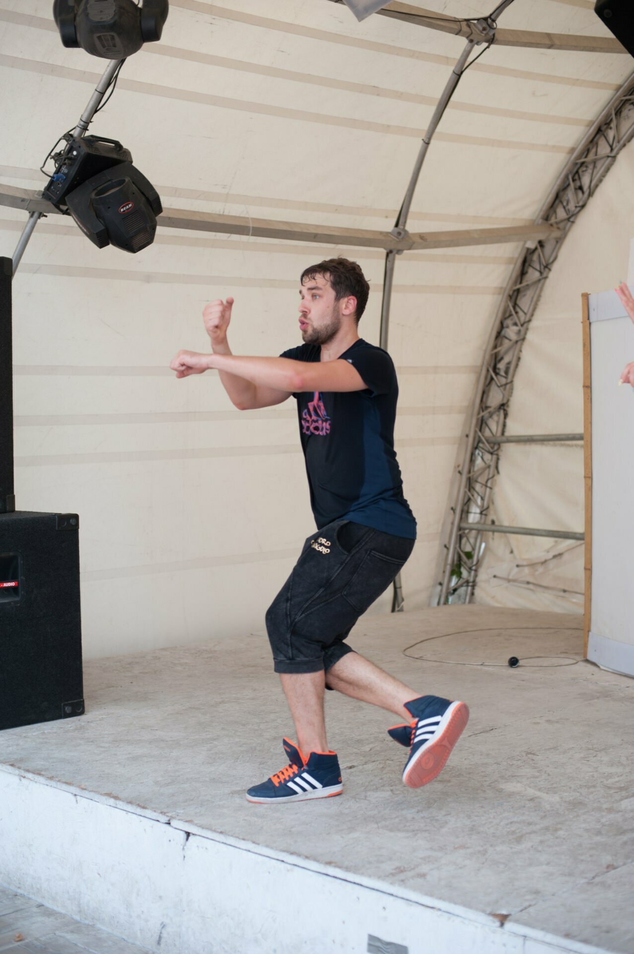 A man in a navy blue shirt and black shorts dances enthusiastically on the stage in the Photo Fair. He is in mid-motion, with his hands in front of him and one leg bent. Speakers and stage equipment are visible in the background. He is wearing blue and orange tennis shoes.   