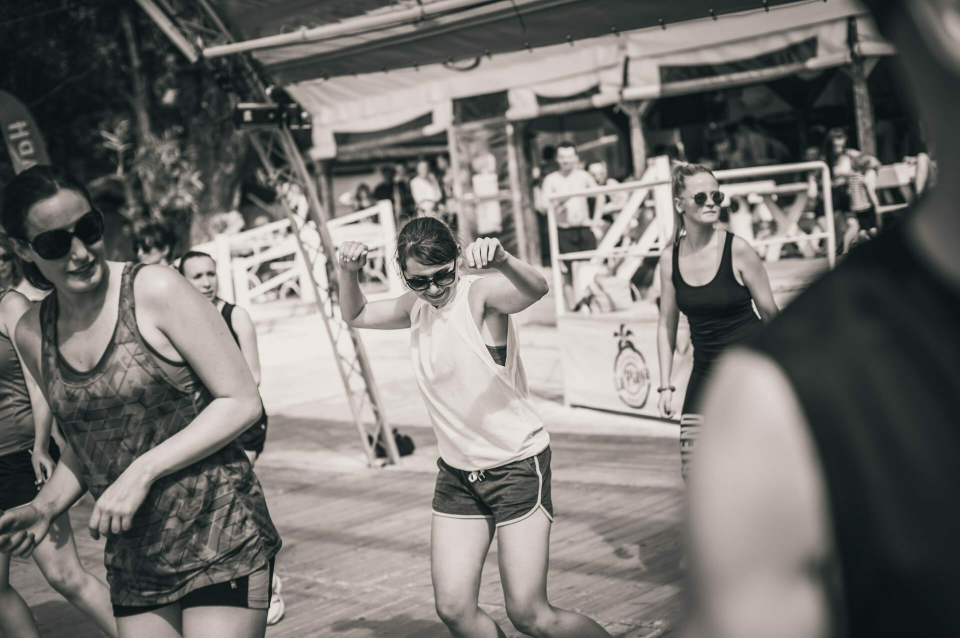 A group of people are dancing outdoors in a lively atmosphere. They are dressed in casual sportswear, and one woman in the middle moves joyfully with her arms raised. In the background is a structure with a shaded area and additional observers, reminiscent of a photo fair.  