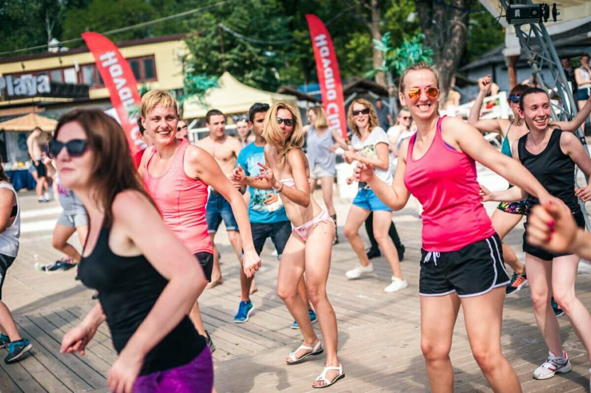 Grupa ludzi, głównie kobiet, energicznie tańczy na drewnianym tarasie na świeżym powietrzu. Większość ma na sobie swobodną odzież sportową, taką jak podkoszulki i szorty. W tle widać czerwone flagi i kilku widzów. Atmosfera wydaje się żywa i radosna, przypominając tętniącą życiem fotorelację z targów.