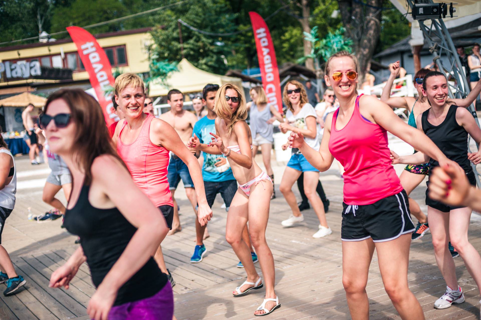 A group of people, mostly women, are vigorously dancing on an outdoor wooden terrace. Most are wearing casual sportswear such as T-shirts and shorts. Red flags and a few spectators can be seen in the background. The atmosphere seems lively and joyful, reminiscent of a bustling photo fair.   