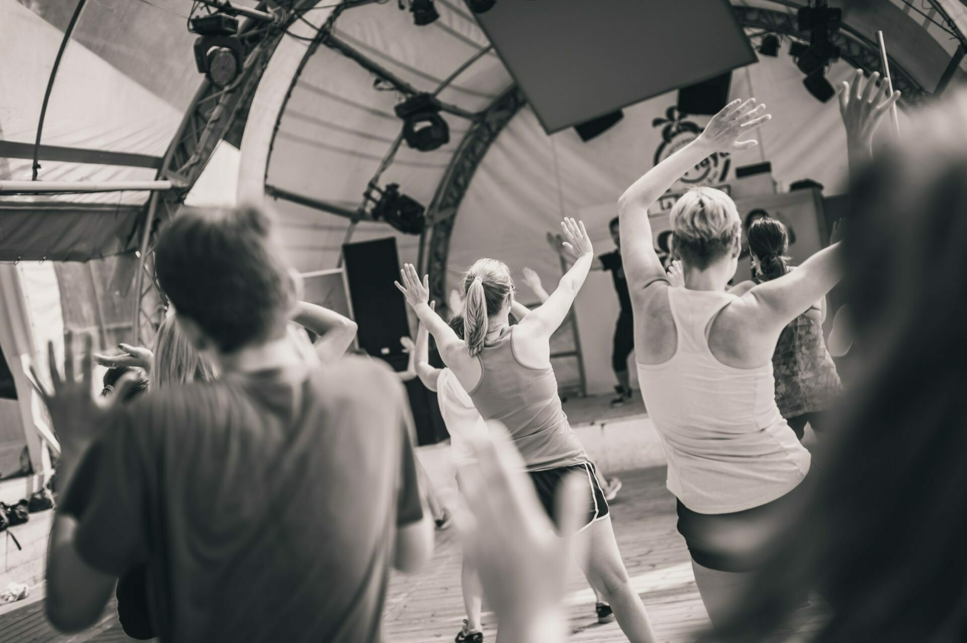 Black and white image of a group of people participating in a dance class or exercise inside a large tent-like structure. They stand with their backs to the camera and raise their hands in the air, following the instructor in front, capturing the lively atmosphere of the photo fair. 