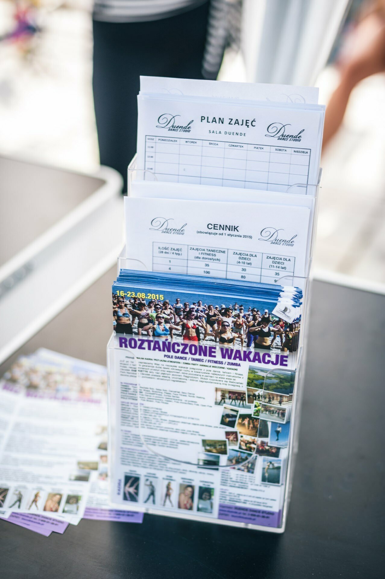 The clear acrylic tabletop brochure rack holds a variety of brochures and schedules. The visible documents bear headings in Polish, including a flyer for the summer dance camp "Roztańczone Wakacje", a schedule "Schedule of Classes", a price list marked "Price List", and a photo report from the fair "Photo Report from the Fair.  