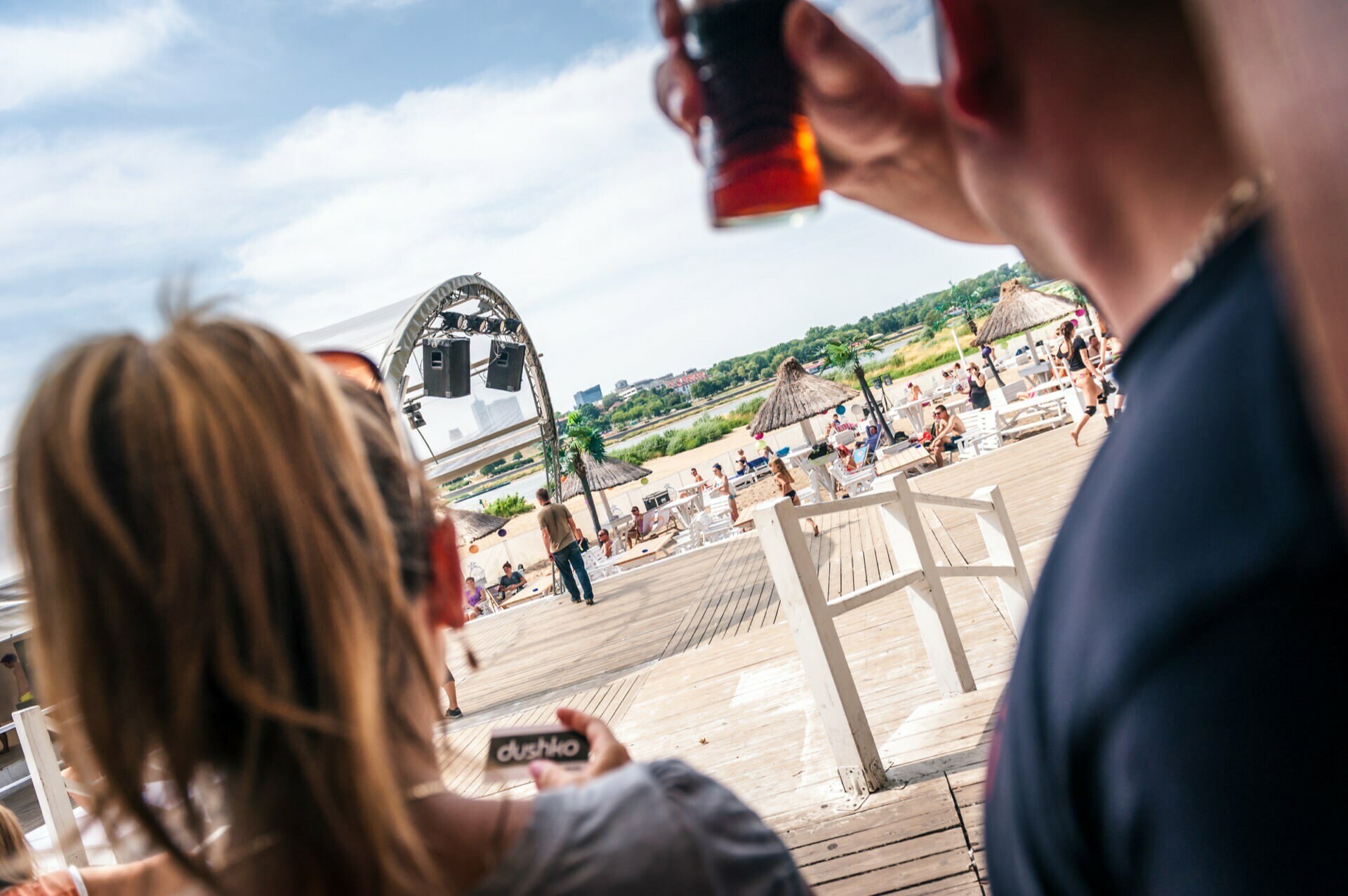 Two people with drinks in their hands are enjoying a sunny day at a beachfront establishment. The area is filled with other people lounging under thatched umbrellas, and the scene is visible in the background. The atmosphere is lively and relaxed, capturing the essence of a photo fair in a seaside setting.  