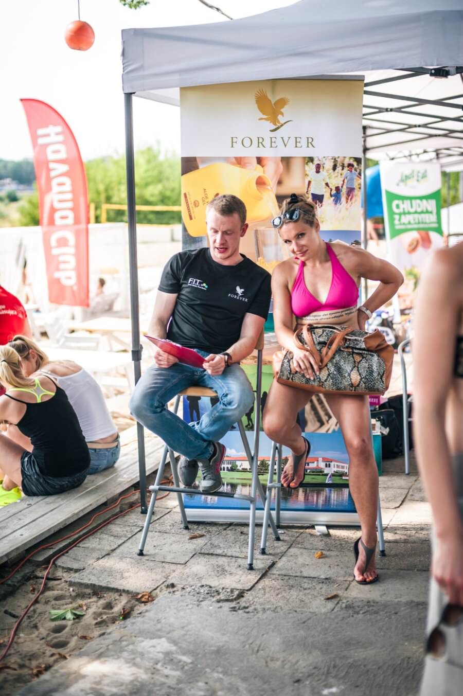 Mężczyzna i kobieta siedzą na wysokich krzesełkach pod baldachimem, a za nimi transparent „Forever”. Mężczyzna w czarnej koszuli trzyma czerwony tablet, a kobieta ubrana w różowy top od bikini i szorty ściska torebkę. Tło wypełniają różnorodne banery i ludzie, tworząc angażującą fotorelację z targów.