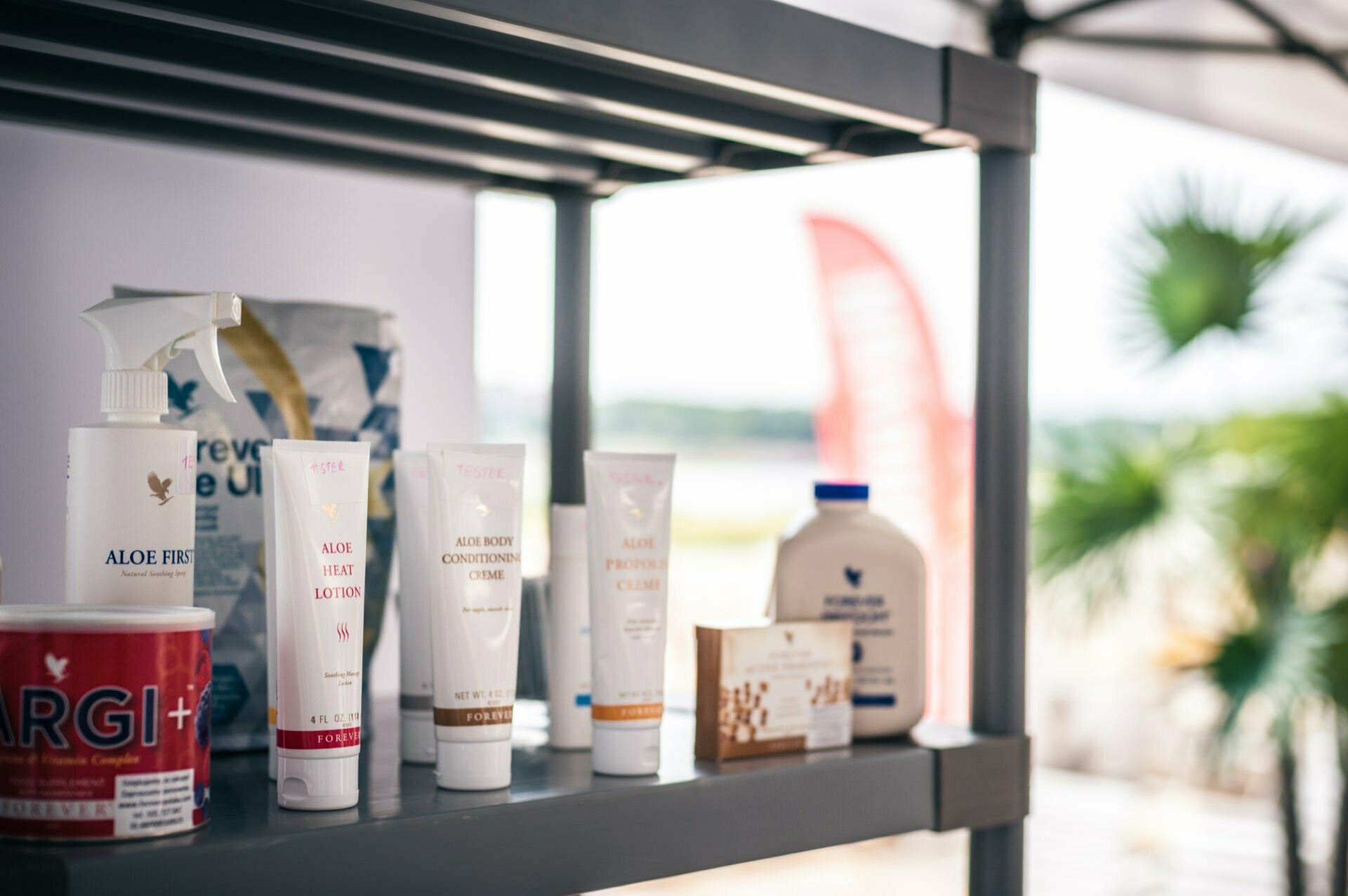 A shelf displaying a variety of beauty and health products, including several bottles and tubes of creams and lotions, a spray bottle, a bag and a tube of dietary supplements. In the background is an outdoor setting with a fuzzy palm tree and a flag, reminiscent of a photo fair. 