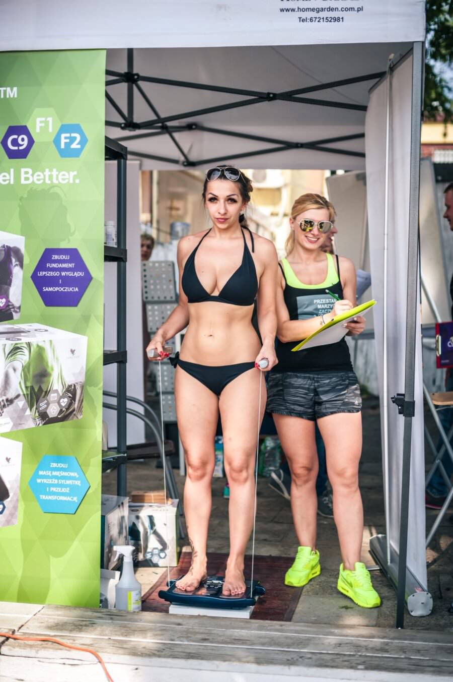 W kabinie znajdują się dwie kobiety, jedna w czarnym bikini stoi boso na urządzeniu. Druga kobieta, w okularach przeciwsłonecznych i swobodnym stroju treningowym, trzyma notes i uśmiecha się. Obok nich na stojaku wisi kolorowy plakat informacyjny, który oddaje istotę fotorelacji z targów.
