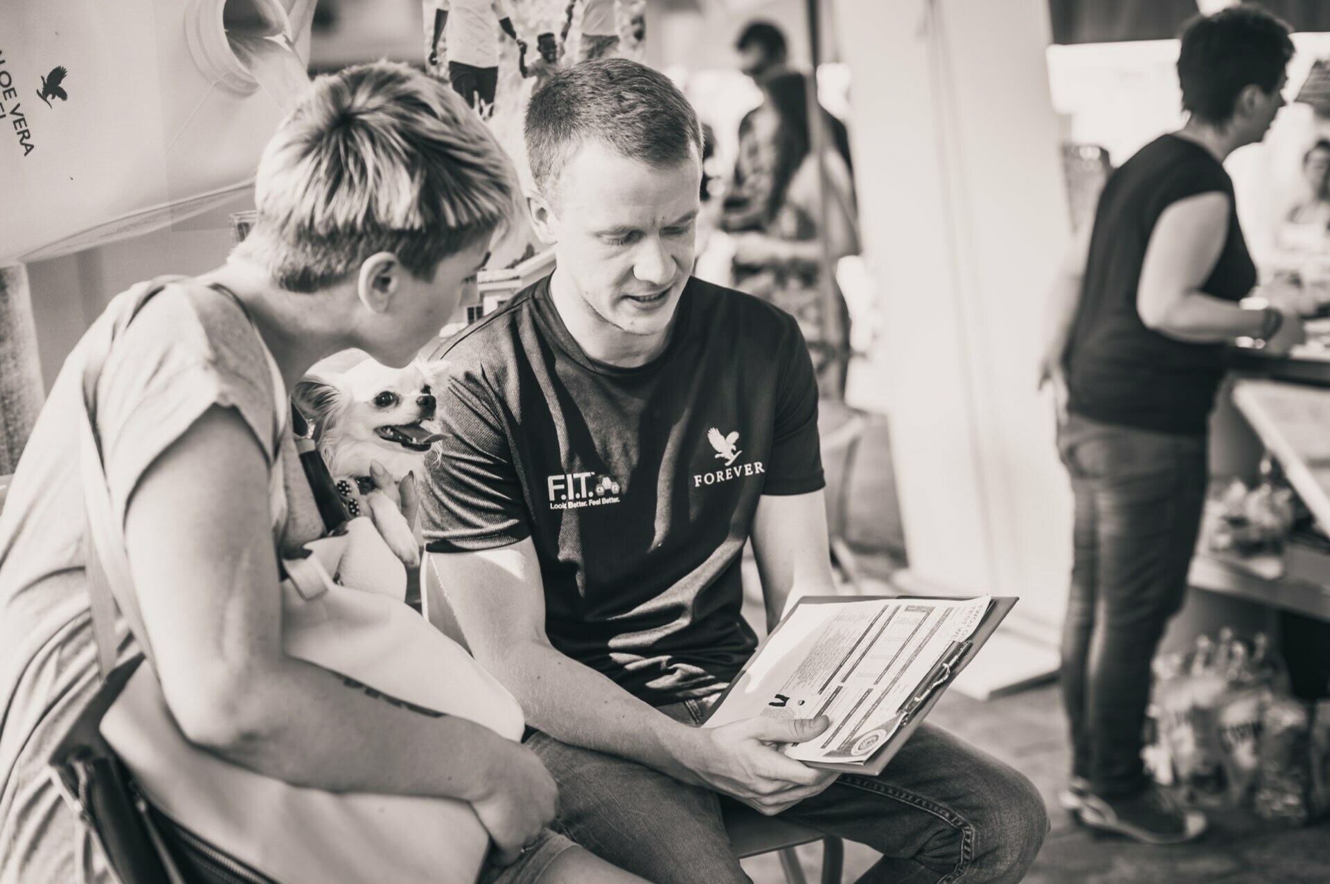 Black and white photo of two people reviewing documents. The person on the left is holding a small dog, the person on the right is wearing a T-shirt that says "Forever." A person can be seen in the background, facing away from the camera, taking a photo report of the fair.  